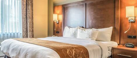 Una habitación de hotel con una cama grande, cabecera de madera, lámparas de noche y una mesita de noche con un reloj.