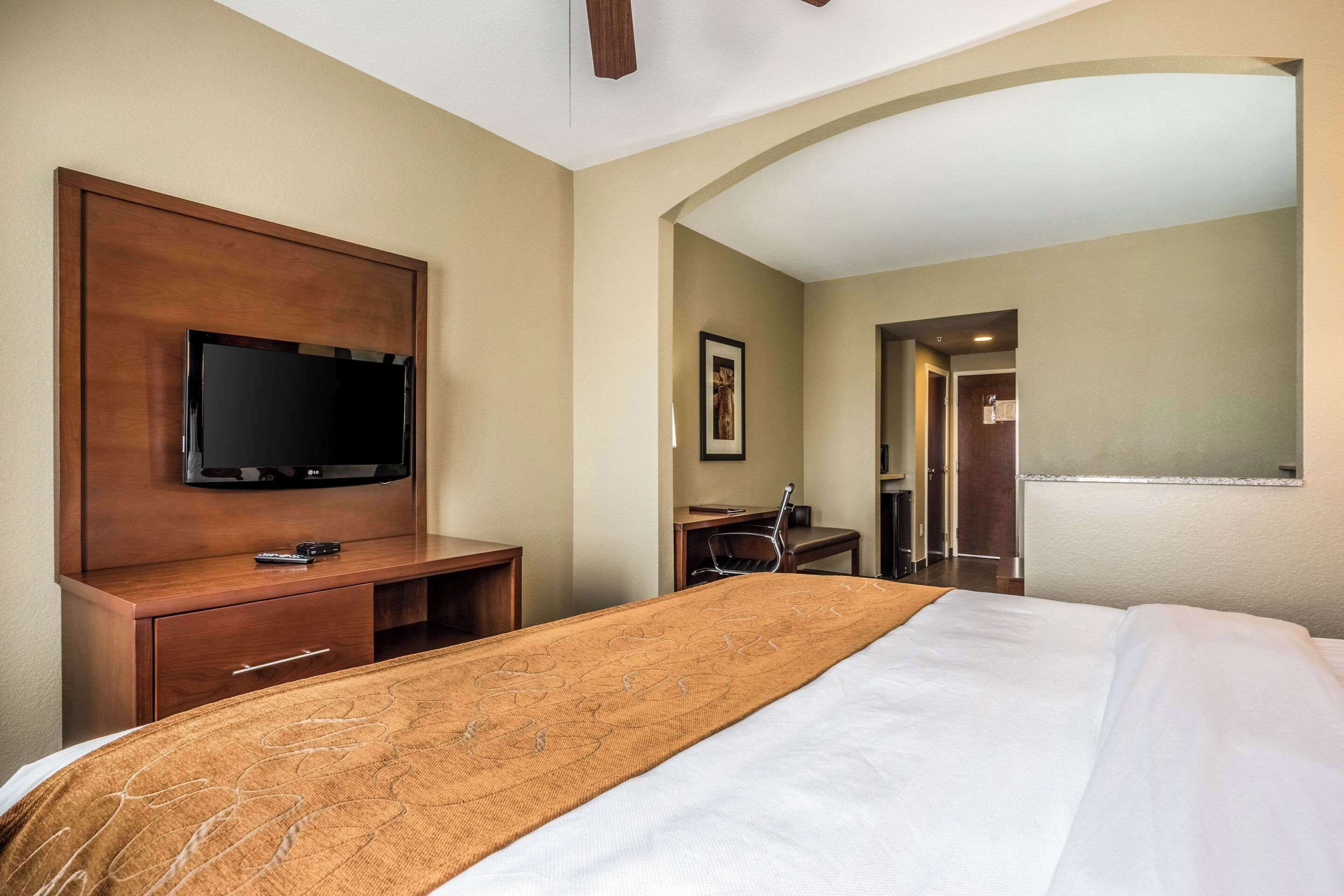 A hotel room with a bed, a TV mounted on a wooden panel, a desk with a chair, and a framed picture on the wall.