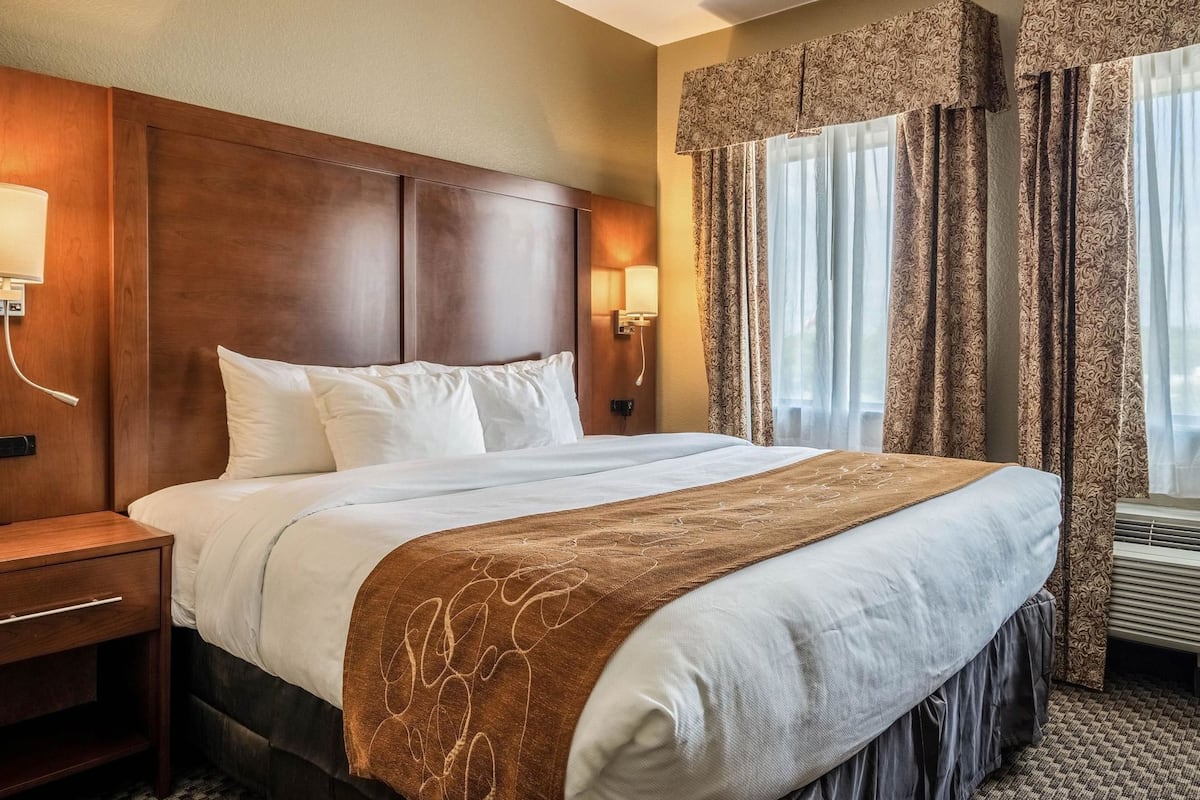Ein Hotelzimmer mit einem großen Bett, einem Holzkopfende, einem Nachttisch, einer Lampe, einer Wandleuchte, gemusterten Vorhängen und einem Fenster mit Klimaanlage.