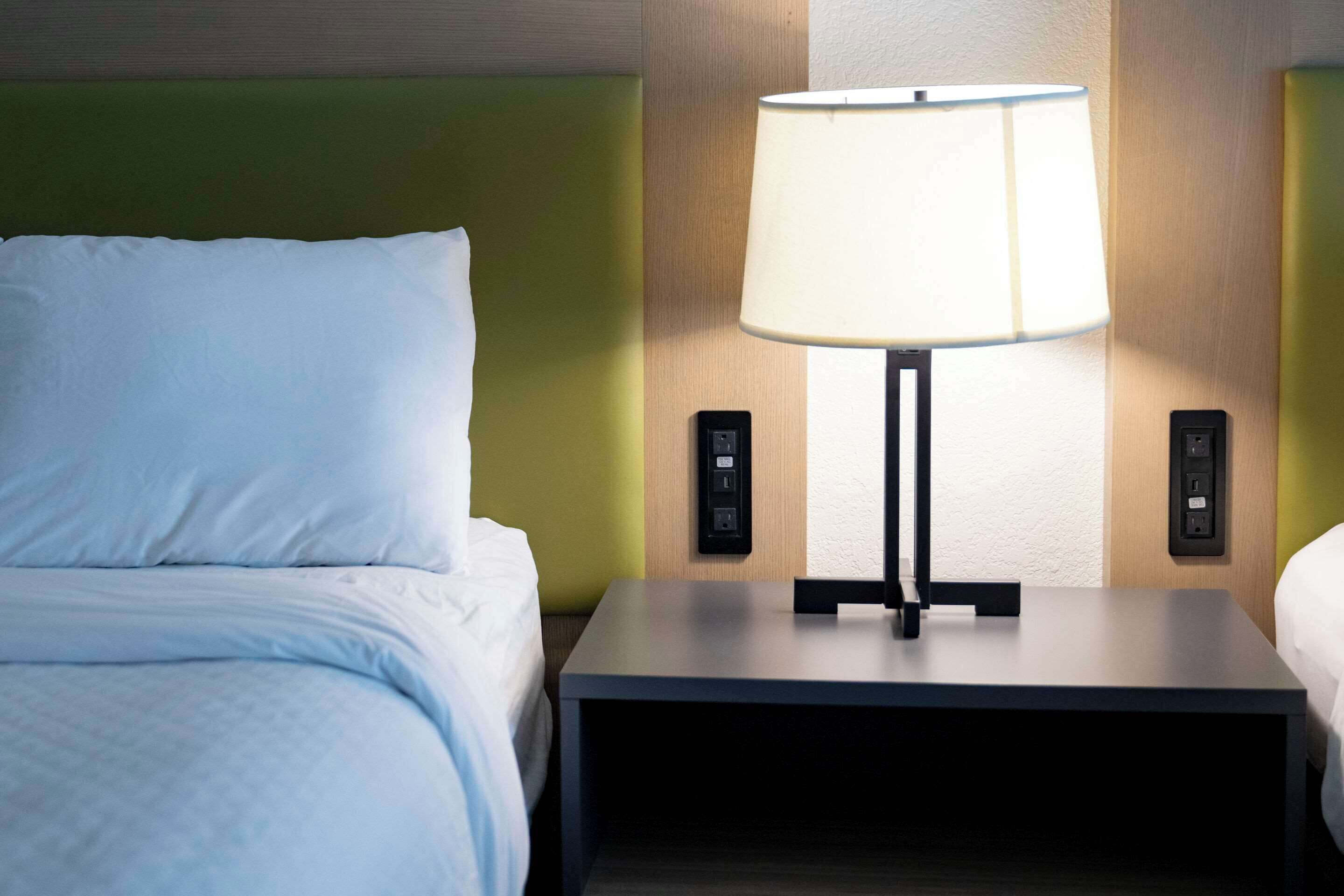 Una cama bien tendida con sábanas blancas, una mesita de noche con lámpara y un enchufe de pared.