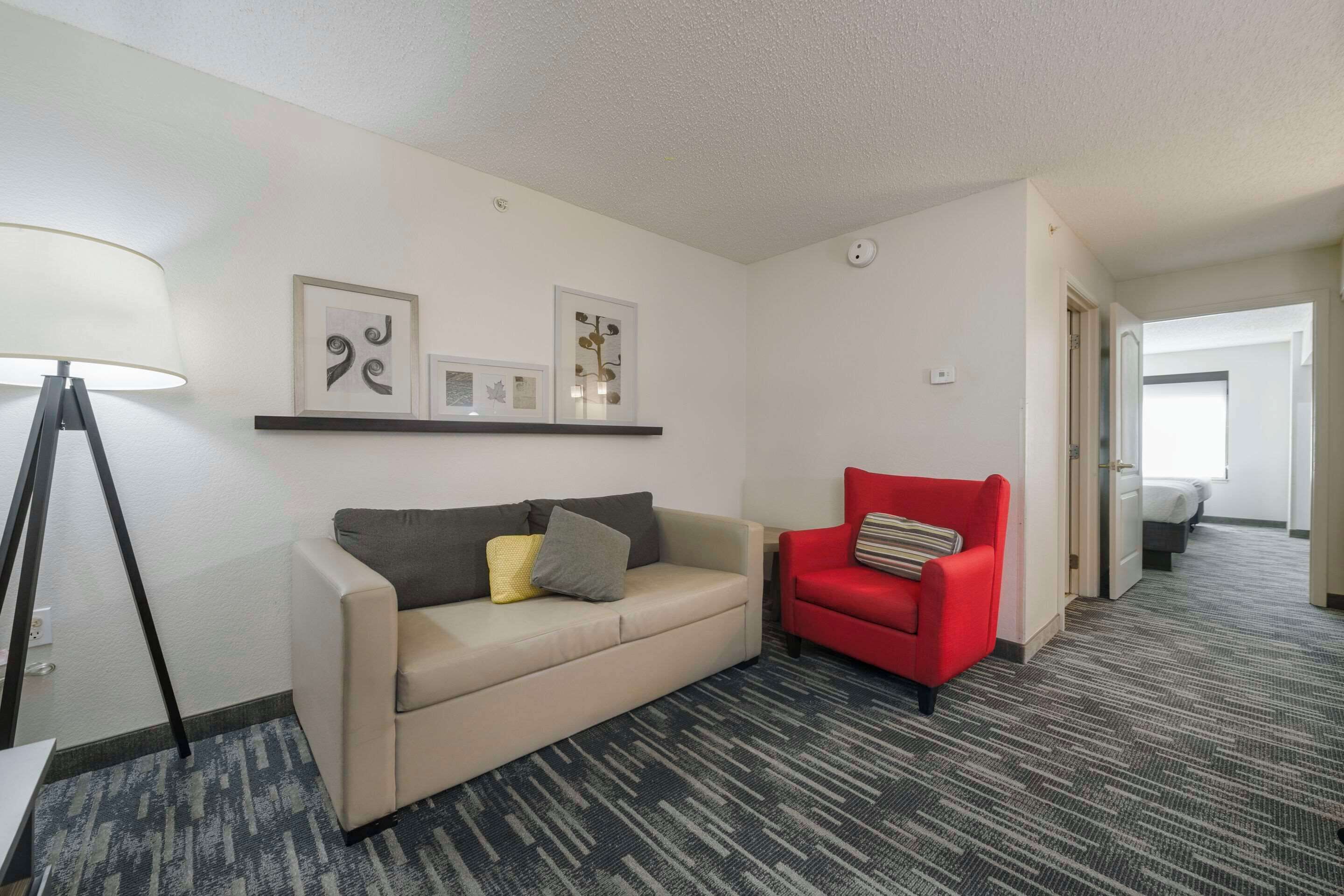A hotel room with a beige sofa, a red armchair, a floor lamp, and framed artwork on the wall.