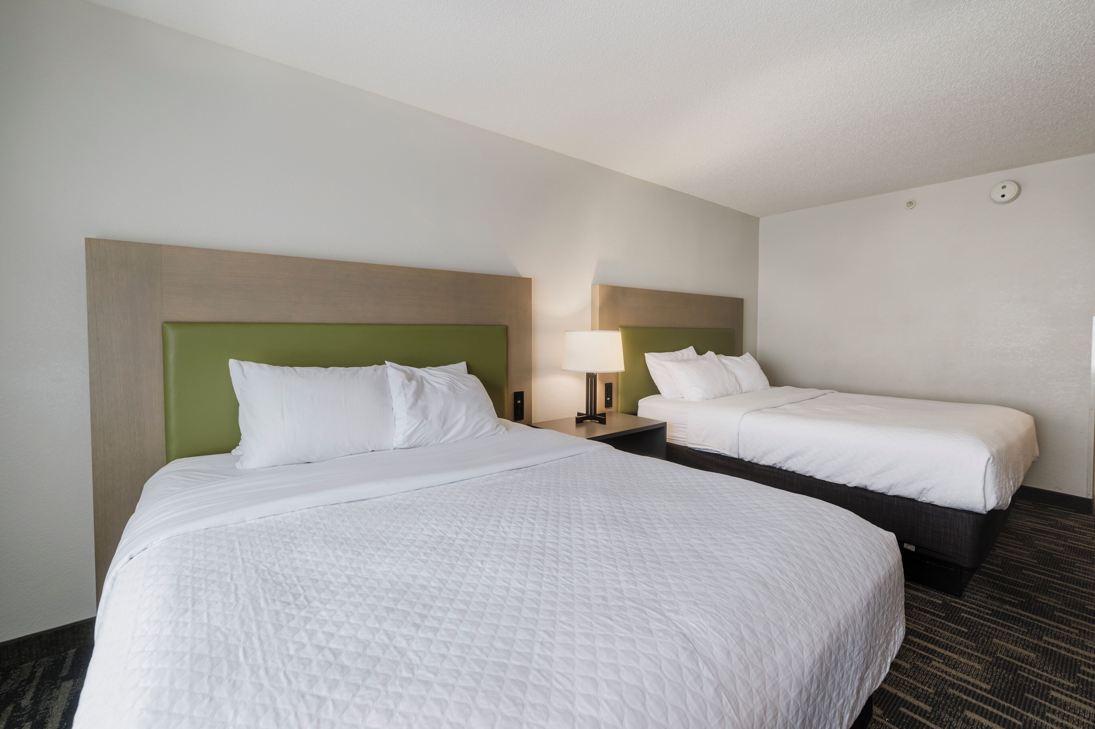 A hotel room with two beds, each with a green headboard, white bedding, and a bedside table with a lamp.