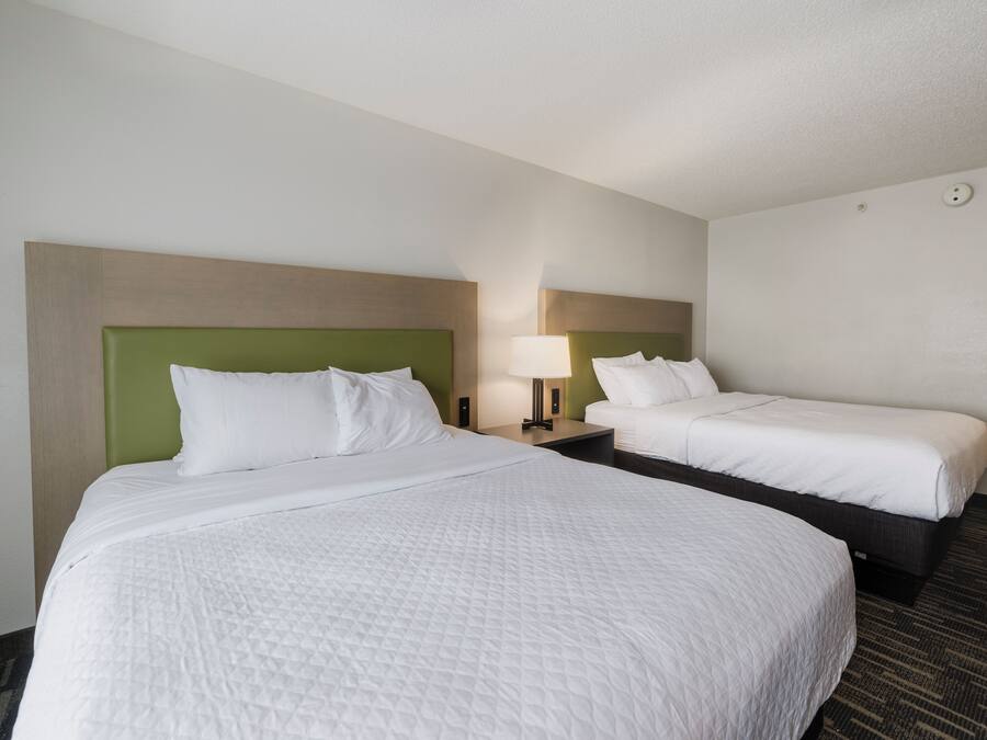 A hotel room with two beds, each with a green headboard, white bedding, and a bedside table with a lamp.