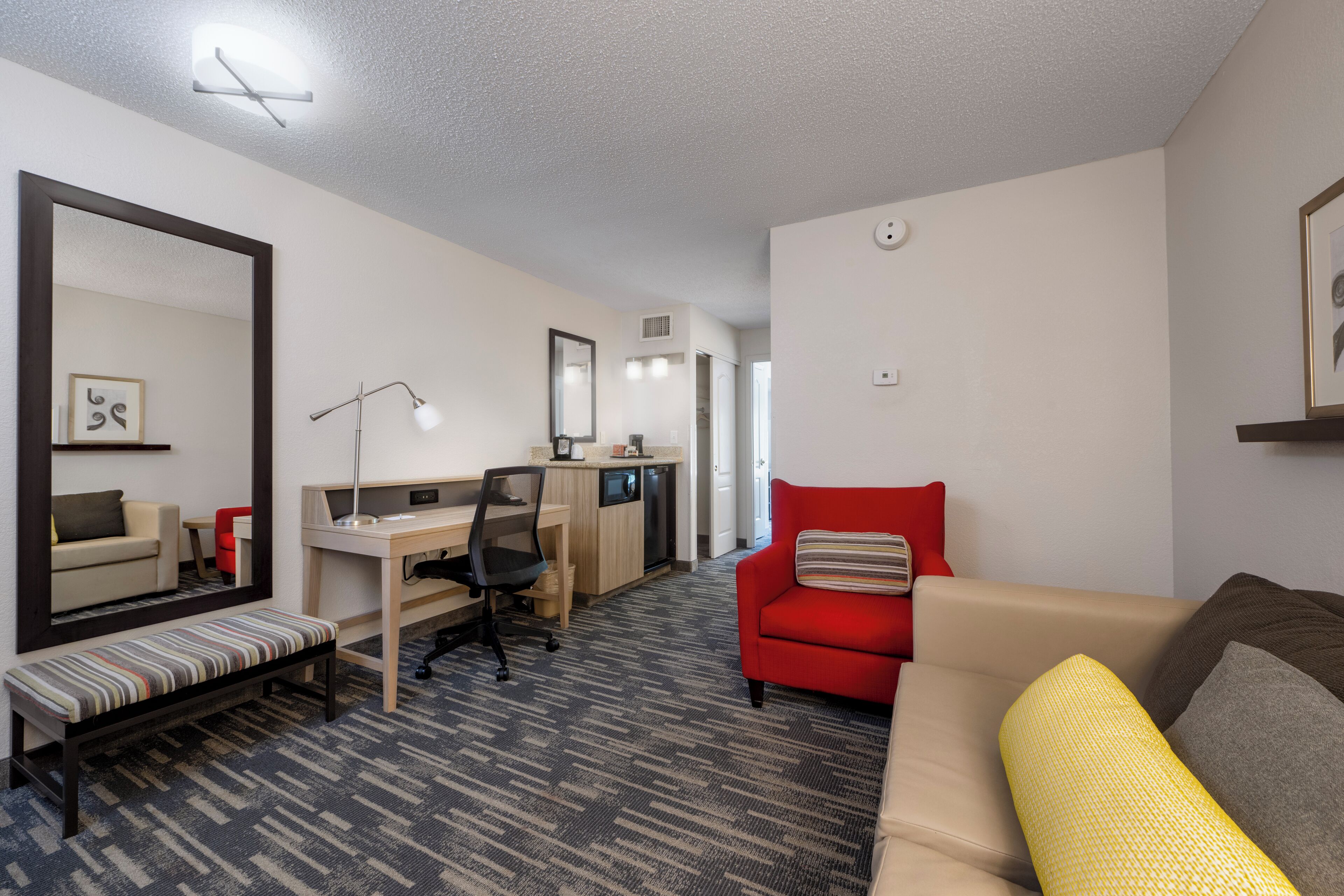 A hotel room with a bed, a desk, a chair, a bench, a red sofa, and a mirror.