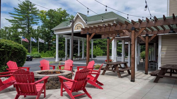 Een terras buiten met rode Adirondack-stoelen, een houten pergola en een overdekt zitgedeelte met picknicktafels.