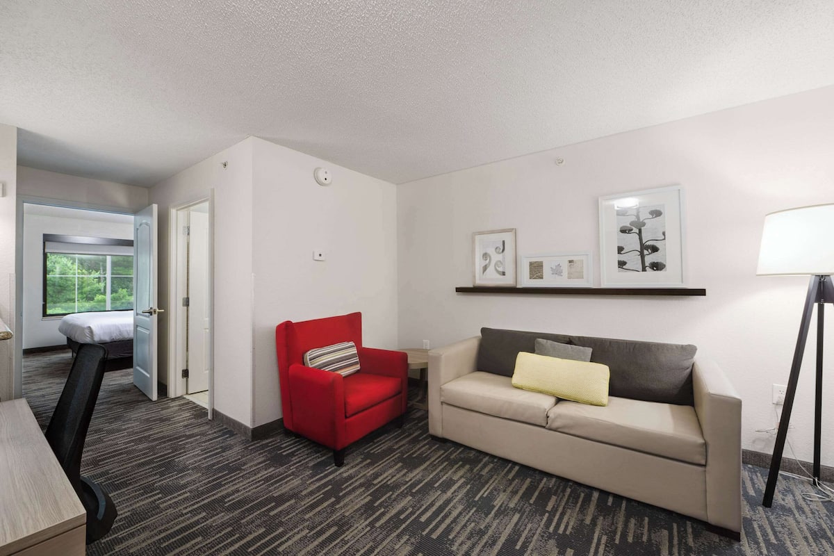 A hotel room with a red armchair, a beige sofa, a small table, and a lamp. There is a window with a view of greenery.