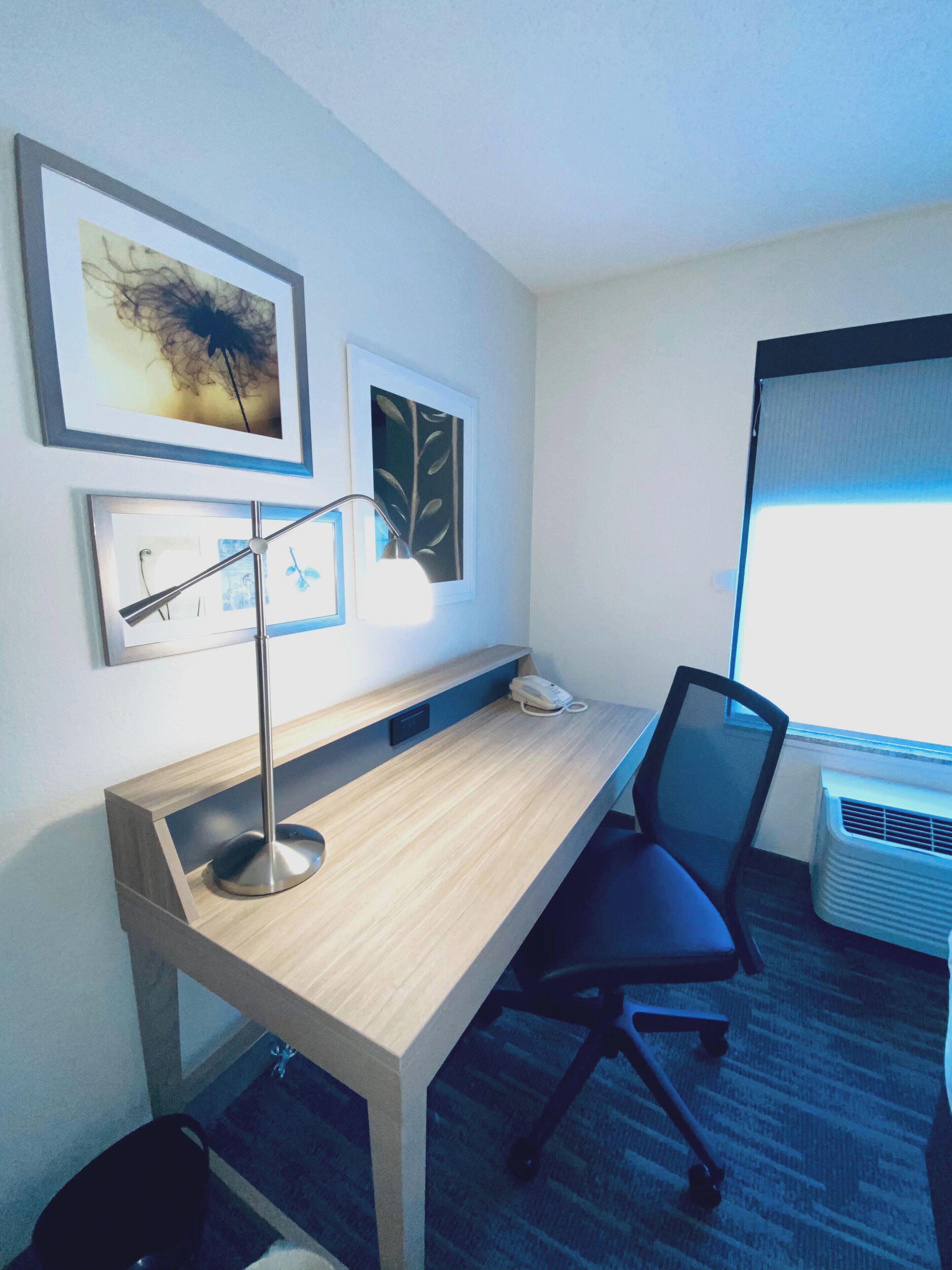 Ein Hotelzimmer mit einem Holztisch, einem Stuhl, einer Schreibtischlampe, einem Telefon und gerahmten Kunstwerken an der Wand.