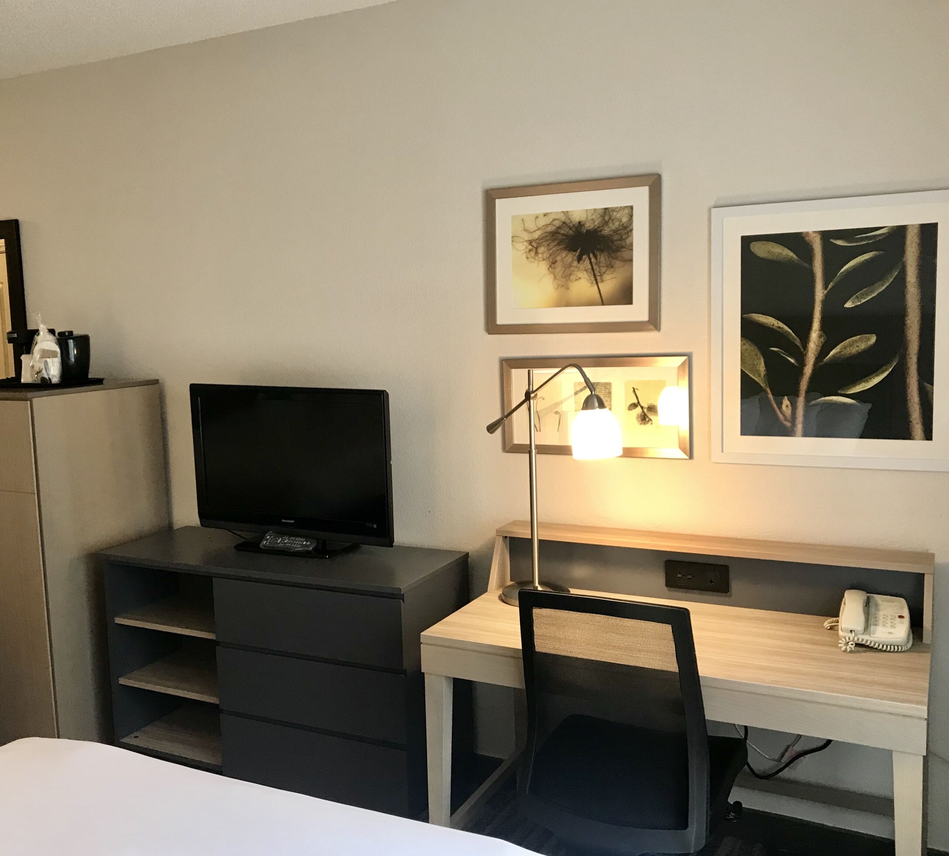 A hotel room with a bed, a desk with a chair, a television, a desk lamp, and two framed pictures on the wall.