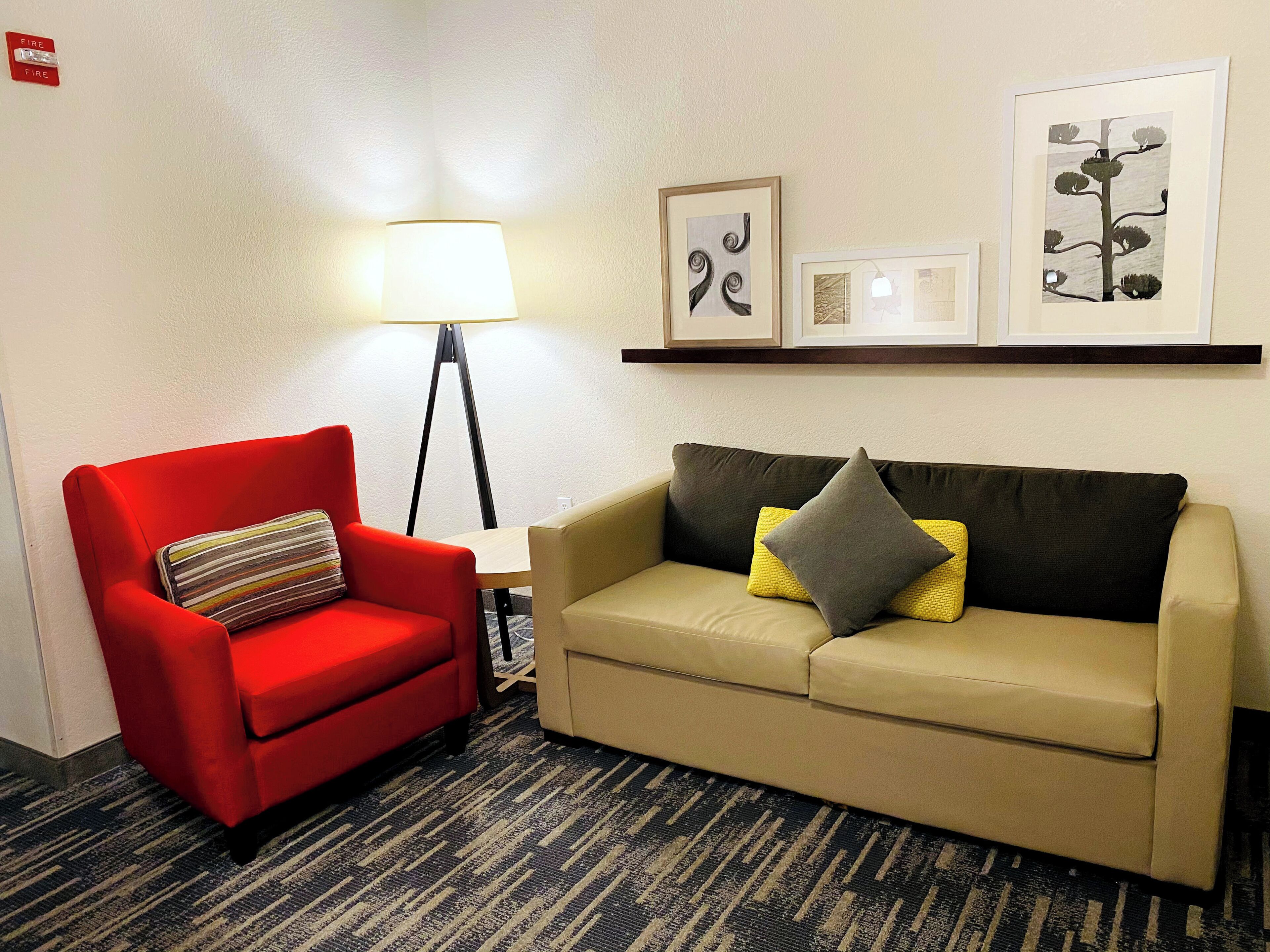 Un sillón rojo con un cojín a rayas, un sofá color beige con dos cojines, una lámpara de pie y obras de arte enmarcadas en la pared.