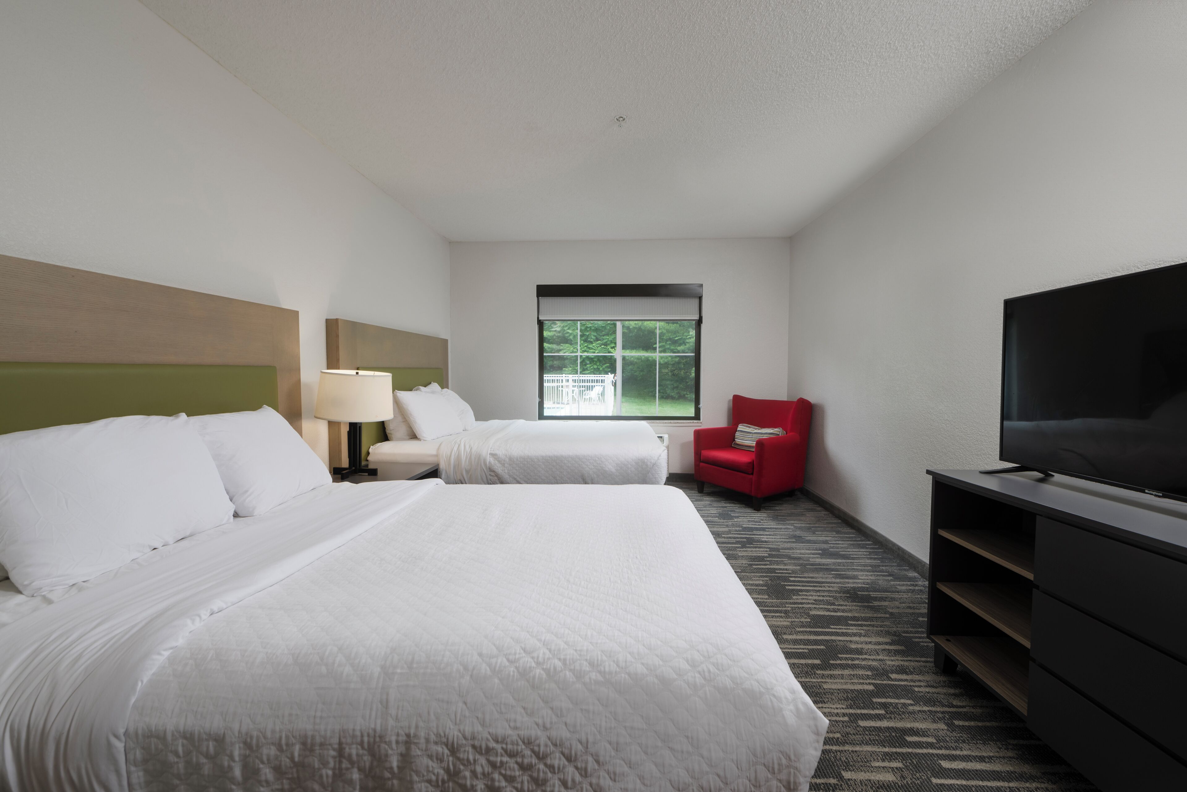 A hotel room with two beds, a TV, a red chair, and a window with a view of greenery.