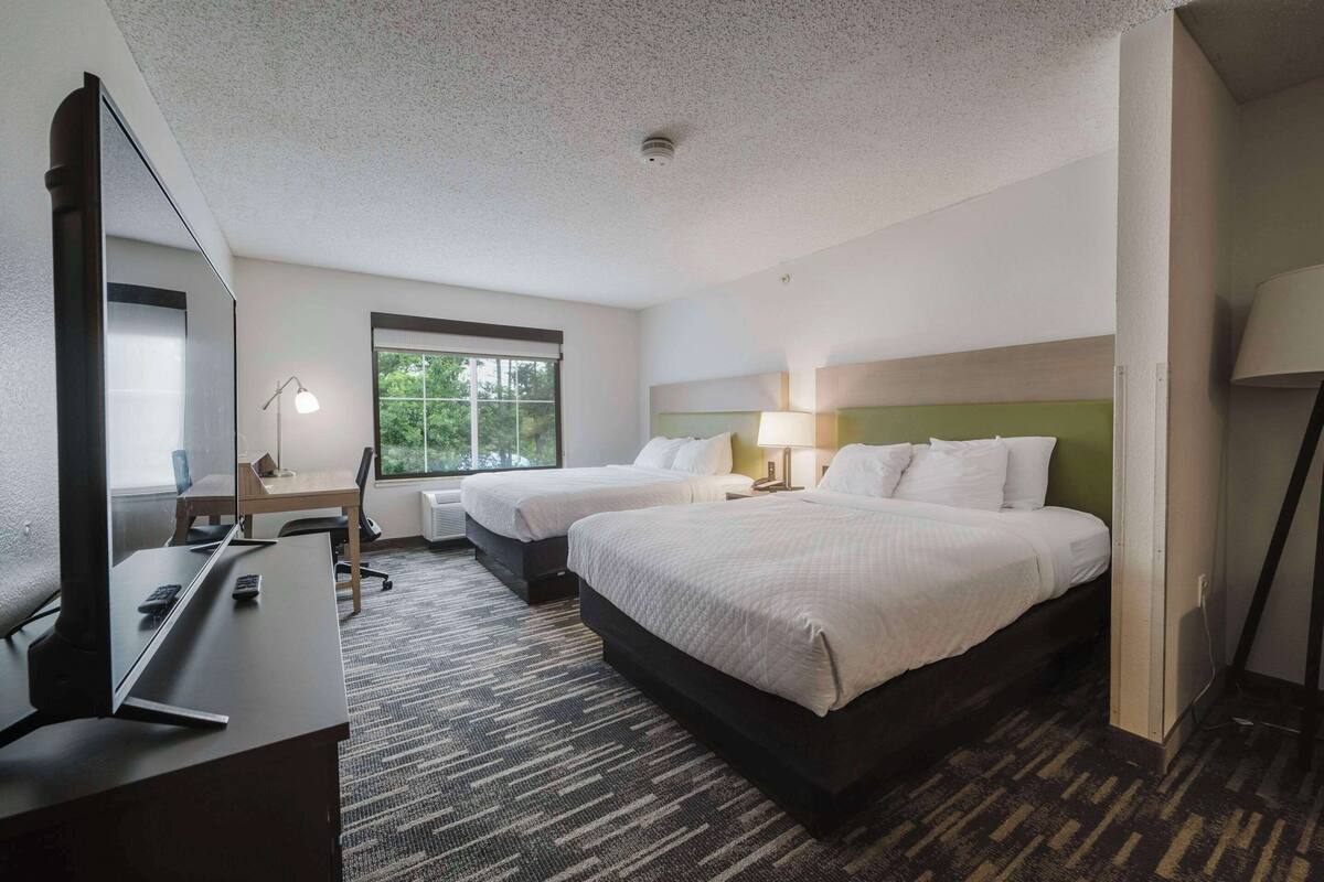 A hotel room with two beds, a desk with a computer, a chair, a lamp, and a window with a view of trees.