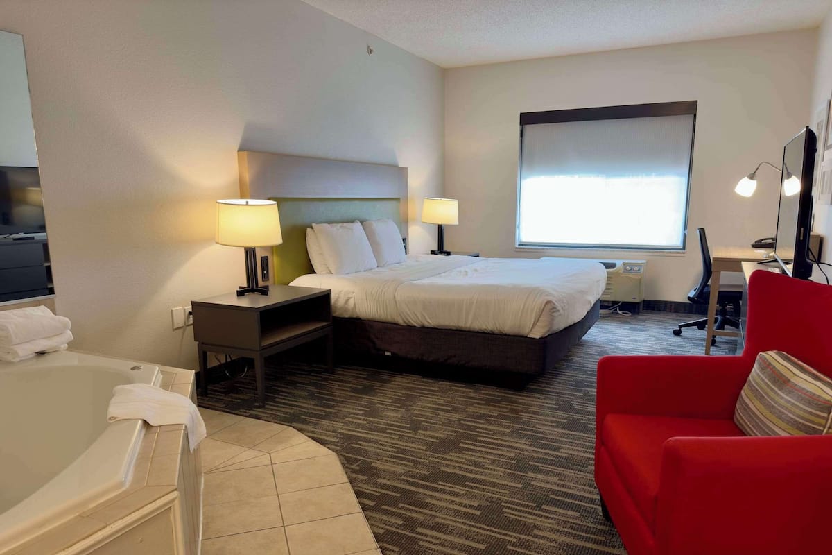 A hotel room with a bed, two bedside lamps, a desk with a computer, a red chair, and a large window.