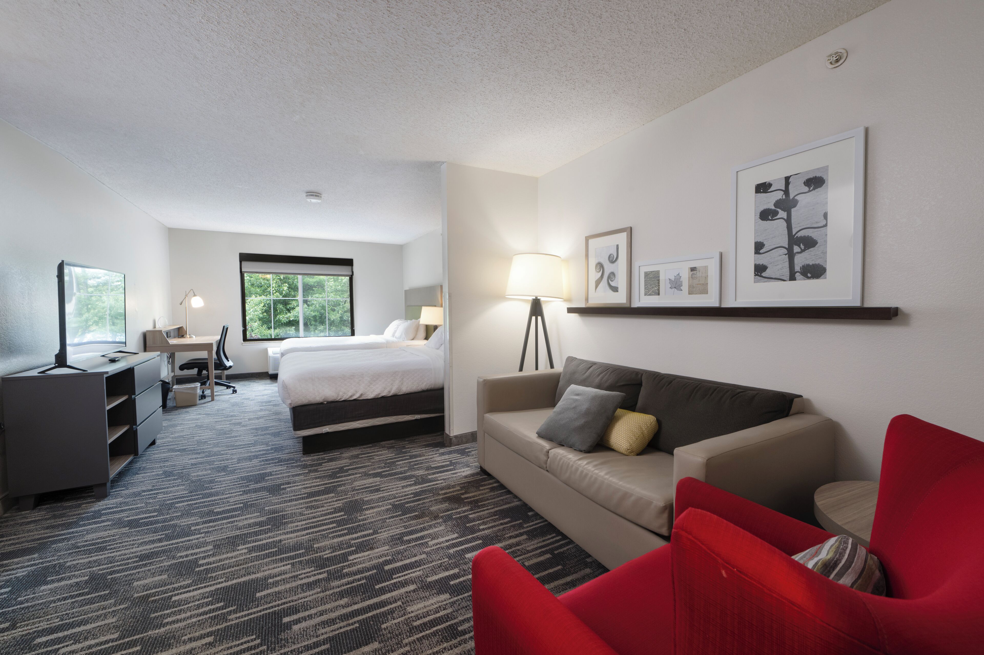 Ein Hotelzimmer mit einem Bett, einem Schreibtisch, einer Couch und einem roten Sessel. An der Wand hängen gerahmte Bilder und auf dem Schreibtisch steht eine Lampe.