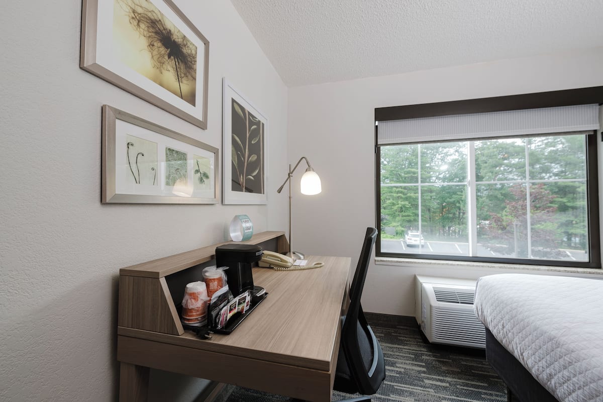 A hotel room with a wooden desk, a lamp, a bed, and a window with a view of trees.