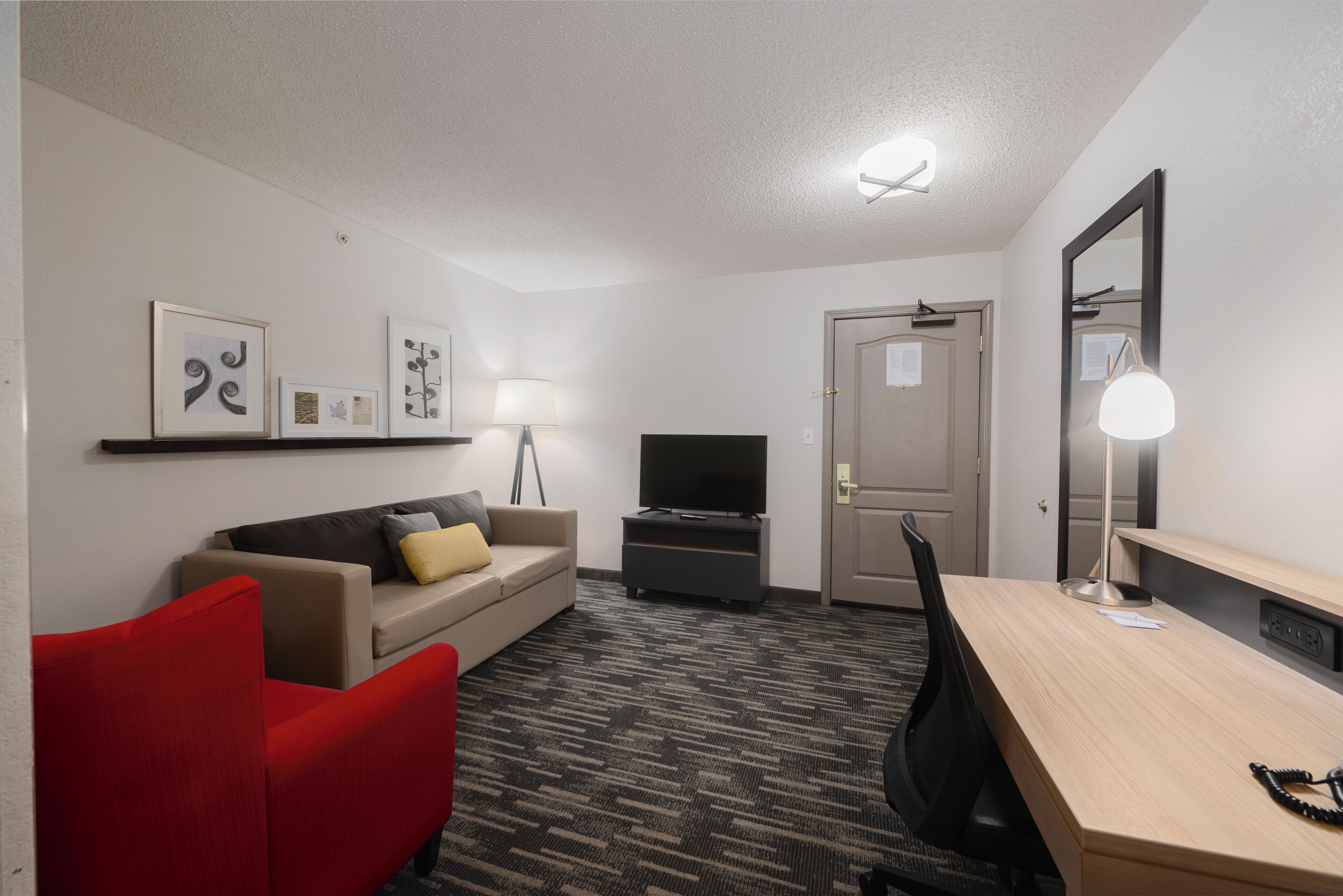 Ein Hotelzimmer mit einer Couch, einem roten Sessel, einem Schreibtisch, einem Fernseher, einer Lampe und gerahmten Kunstwerken an der Wand.