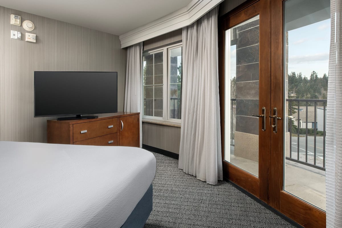 A hotel room with a large bed, a flat-screen TV, a wooden dresser, a window with curtains, and a balcony with a view of trees and a road.