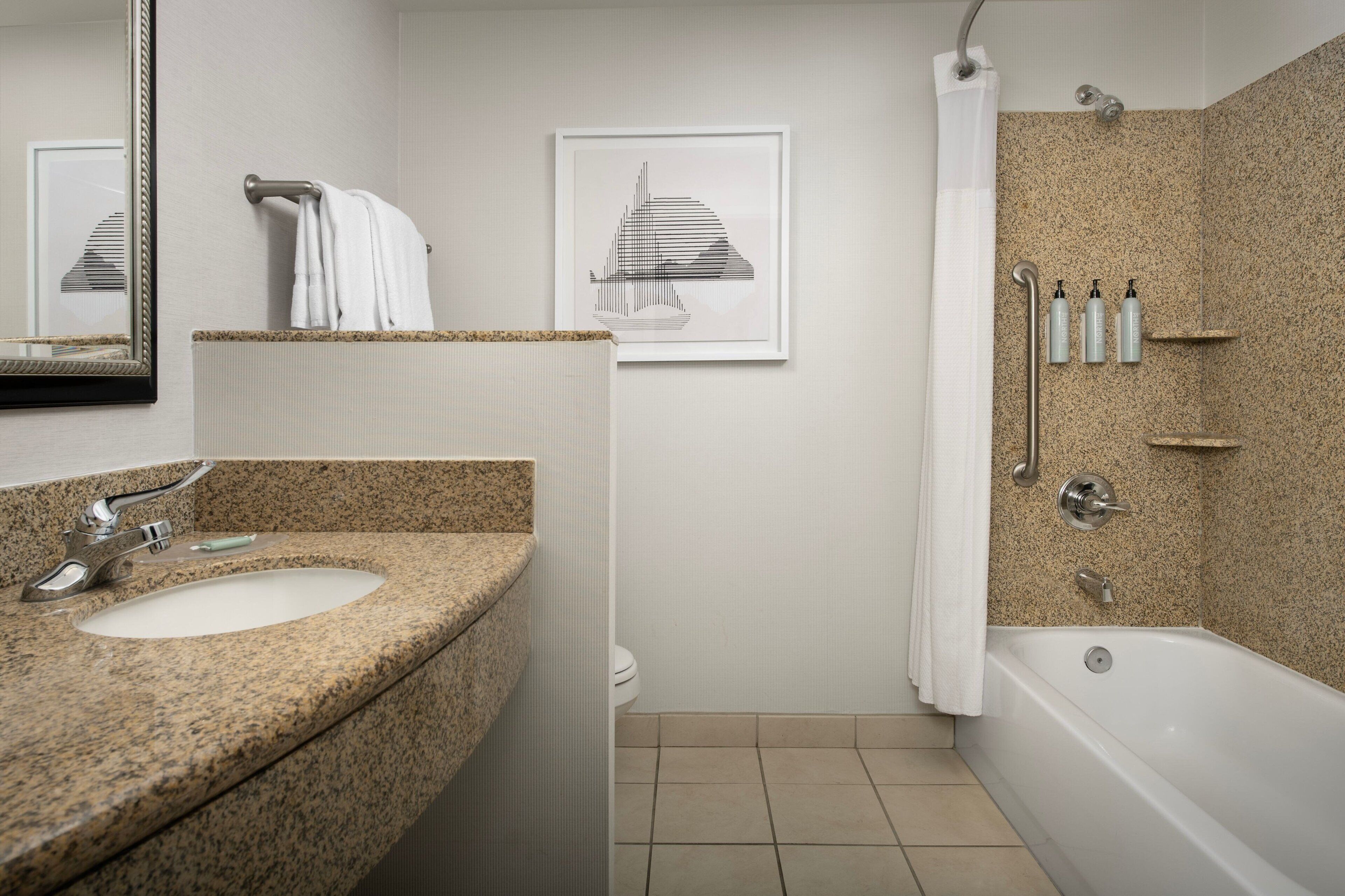 A bathroom with a granite countertop sink, a white toilet, a bathtub, and a shower with a curtain. There are framed pictures on the wall and toiletries on a shelf.