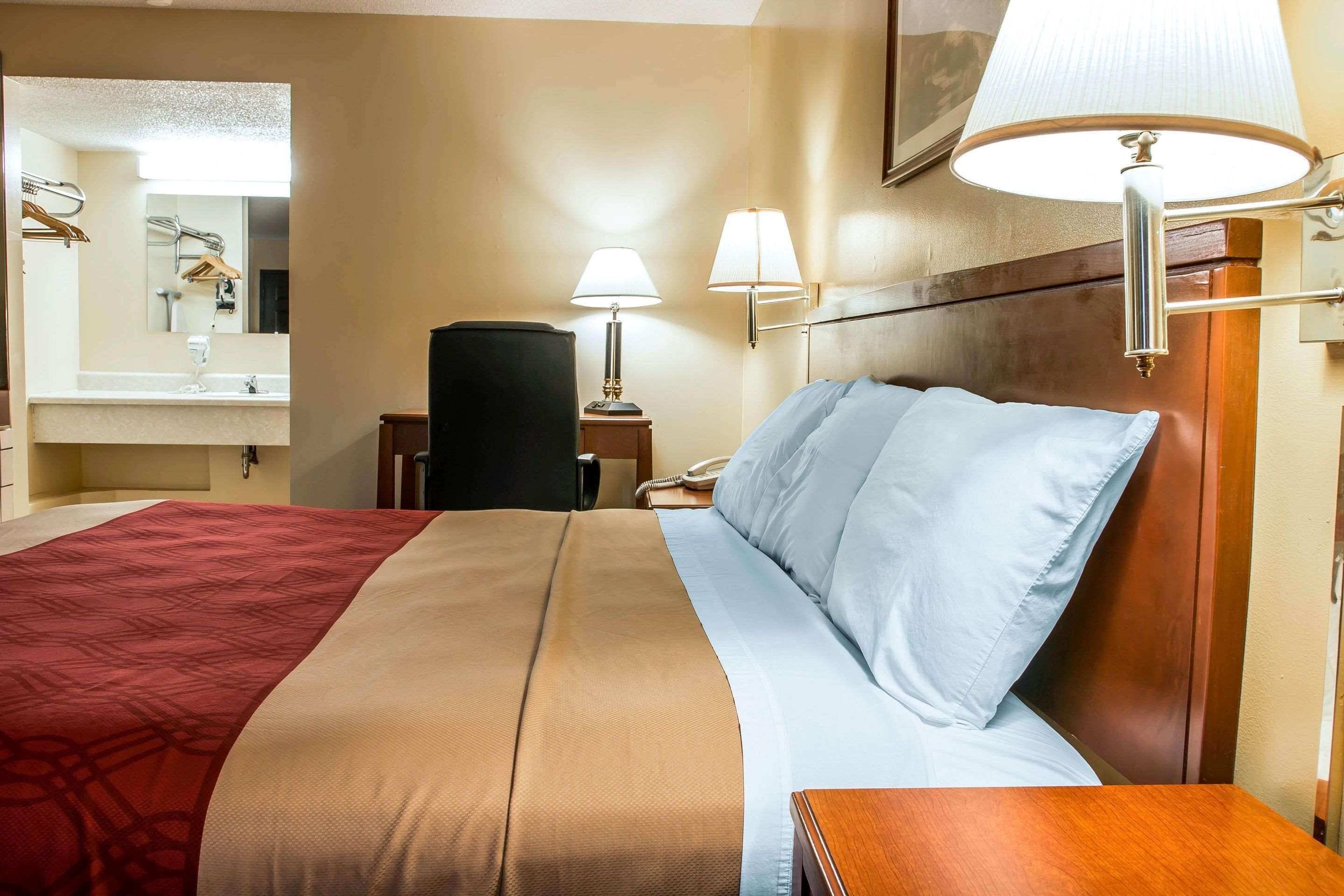 Une chambre d’hôtel avec un grand lit, une tête de lit en bois, une table de chevet avec une lampe, un miroir et une salle de bain équipée d’un lavabo et d’un porte-serviettes.