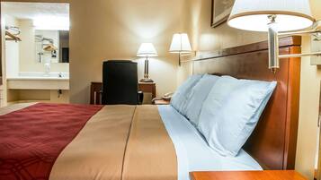 Une chambre d’hôtel avec un grand lit, une tête de lit en bois, une table de chevet avec une lampe, un miroir et une salle de bain équipée d’un lavabo et d’un porte-serviettes.