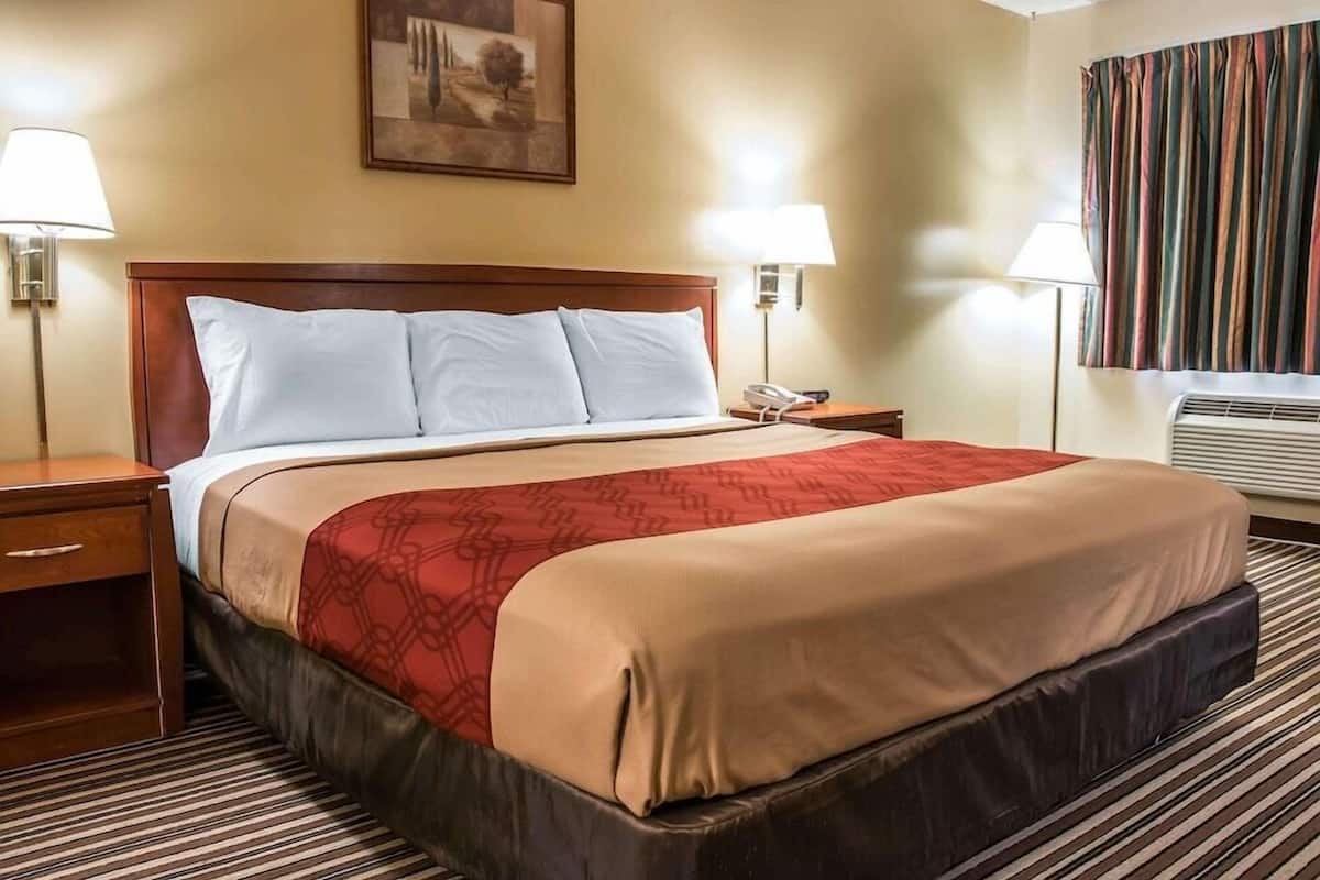 Ein Hotelzimmer mit einem großen Bett, zwei Nachttischen, einem Schreibtisch, einer Wandlampe, einem Telefon und einem Fenster mit Vorhängen.