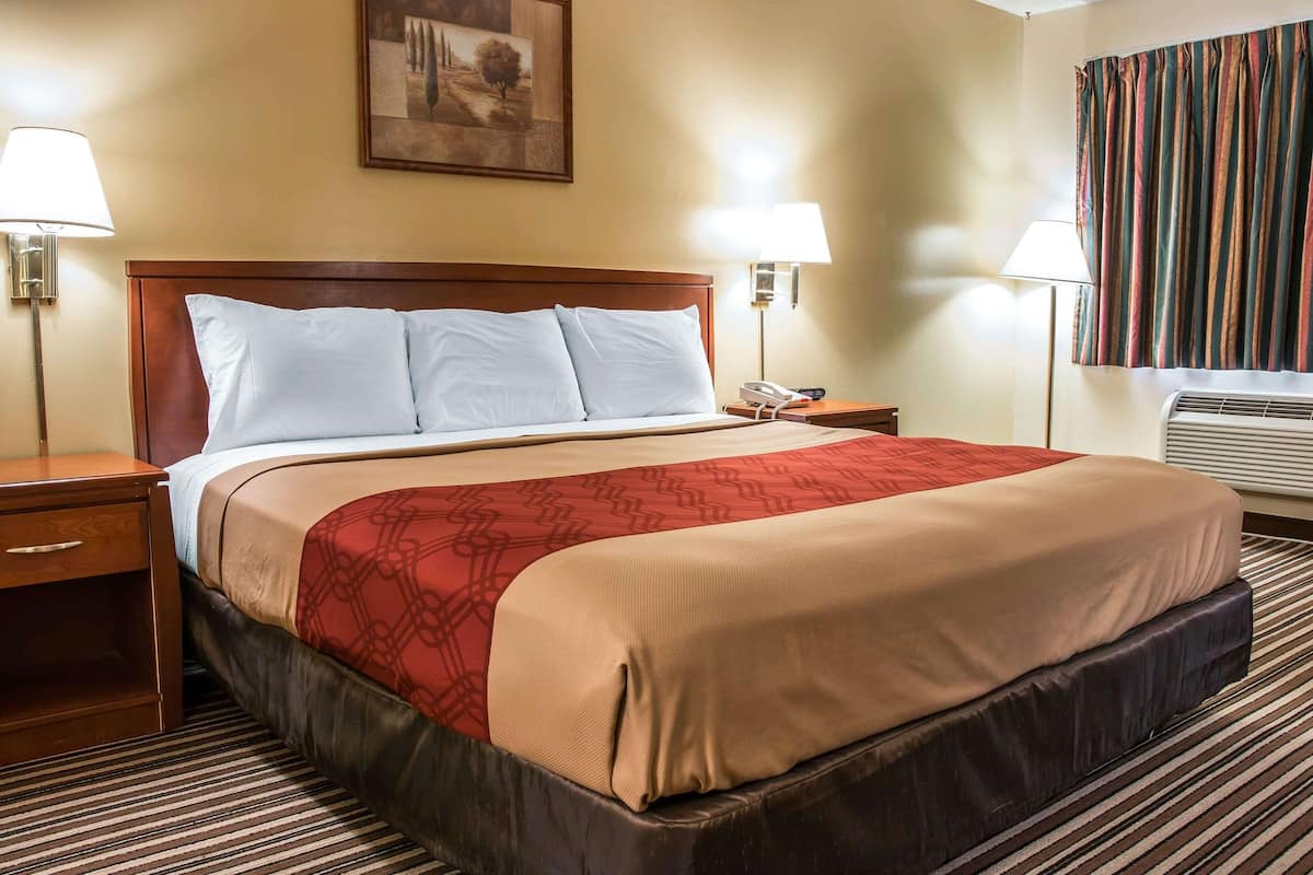 Ein Hotelzimmer mit einem großen Bett, zwei Nachttischen, einem kleinen Tisch, einem an der Wand montierten Telefon und einem Fenster mit Vorhängen.