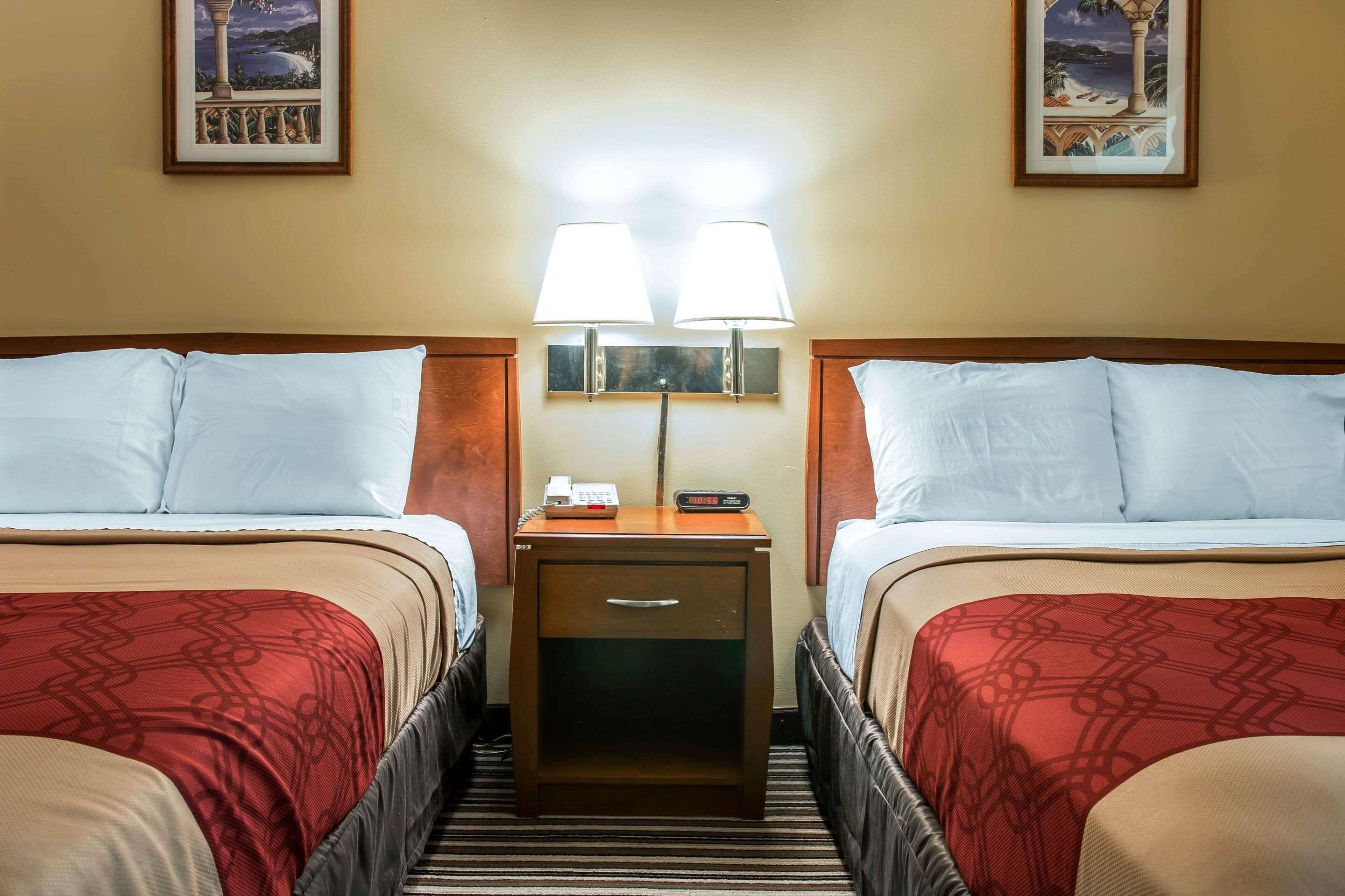Ein Hotelzimmer mit zwei Betten, jeweils mit einer roten, gemusterten Tagesdecke, einem Nachttisch mit Lampe und einer digitalen Uhr.