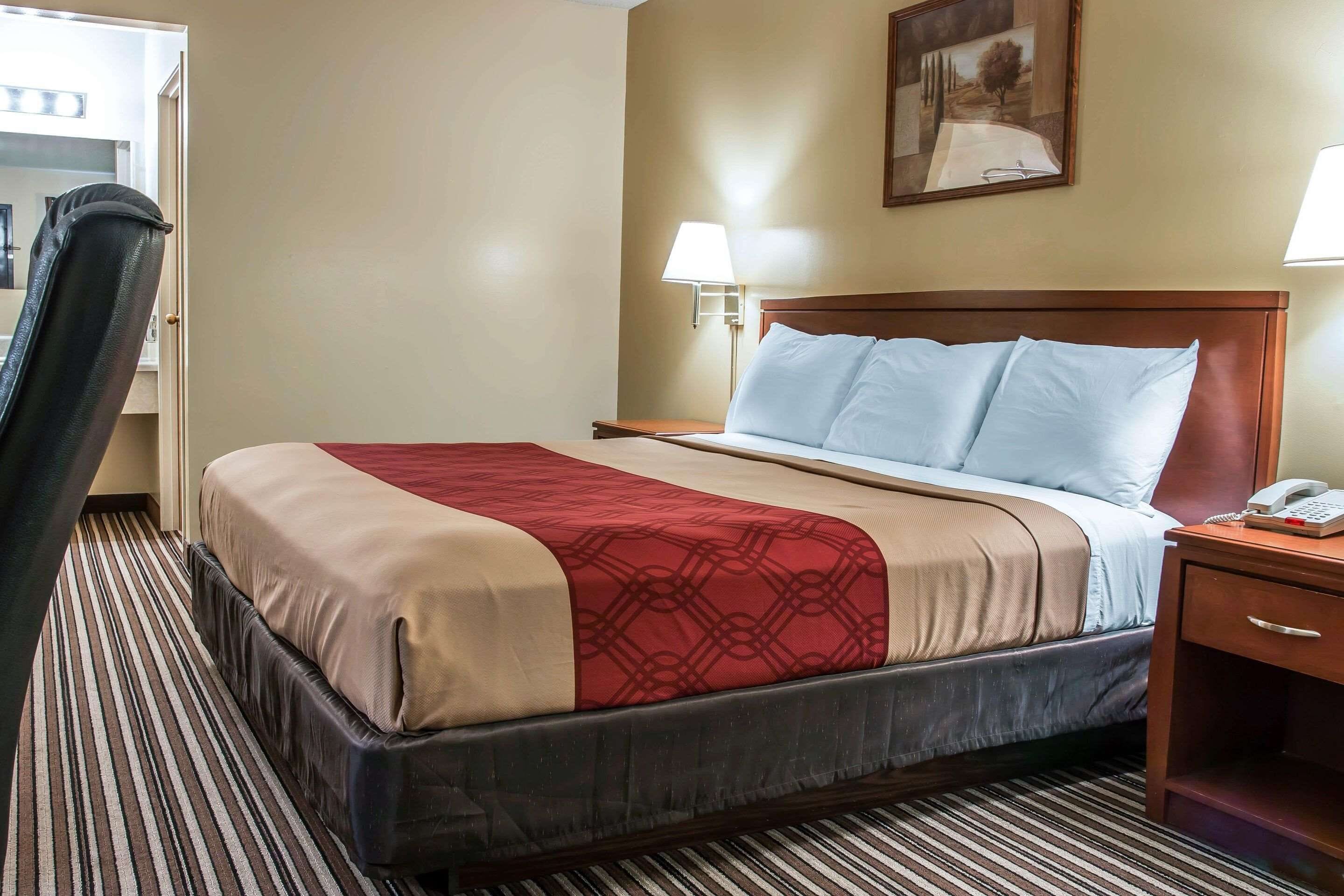 A hotel room with a bed, a bedside table with a lamp, a chair, and a painting on the wall.