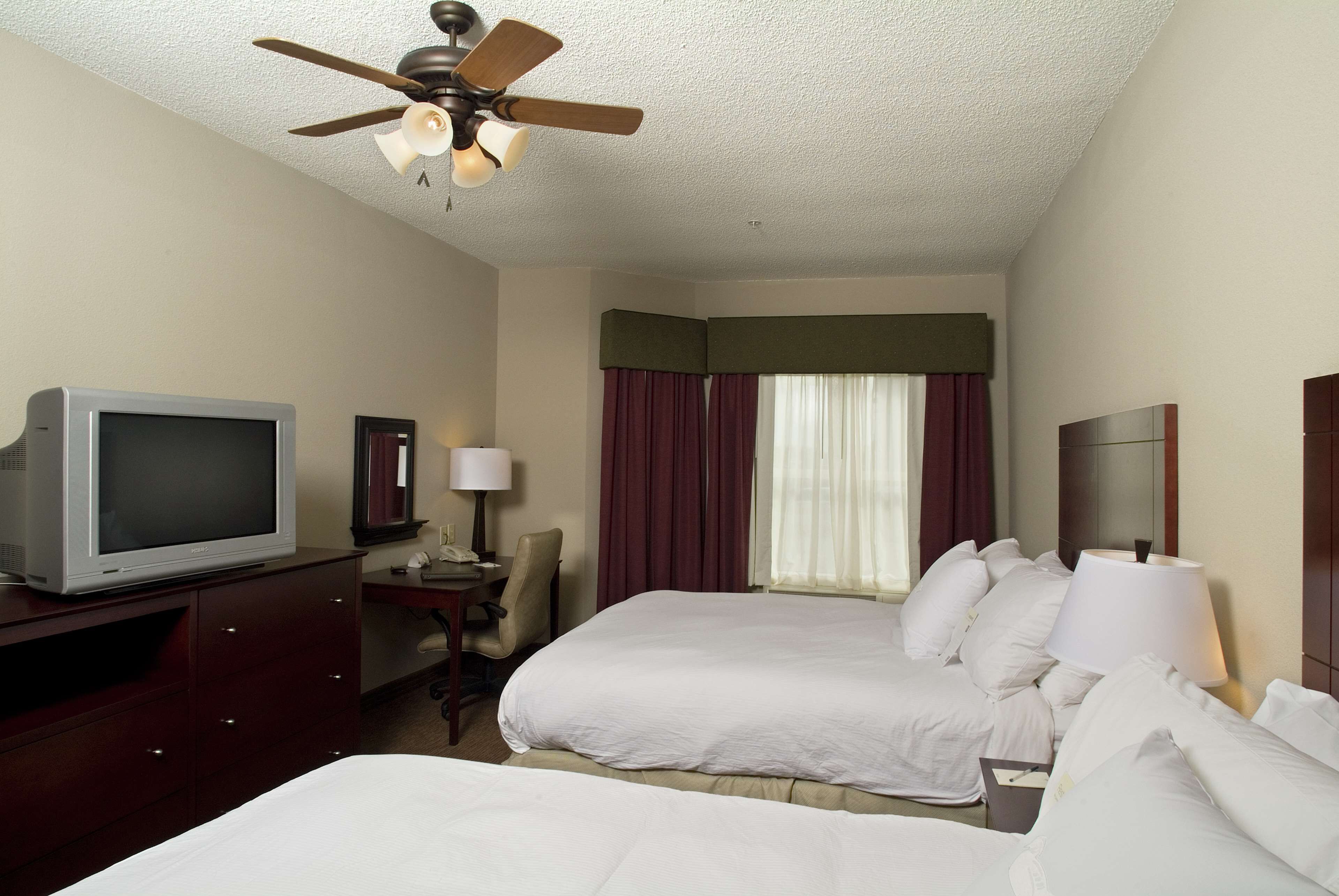 A hotel room with two beds, a television, a desk with a lamp, and a ceiling fan.
