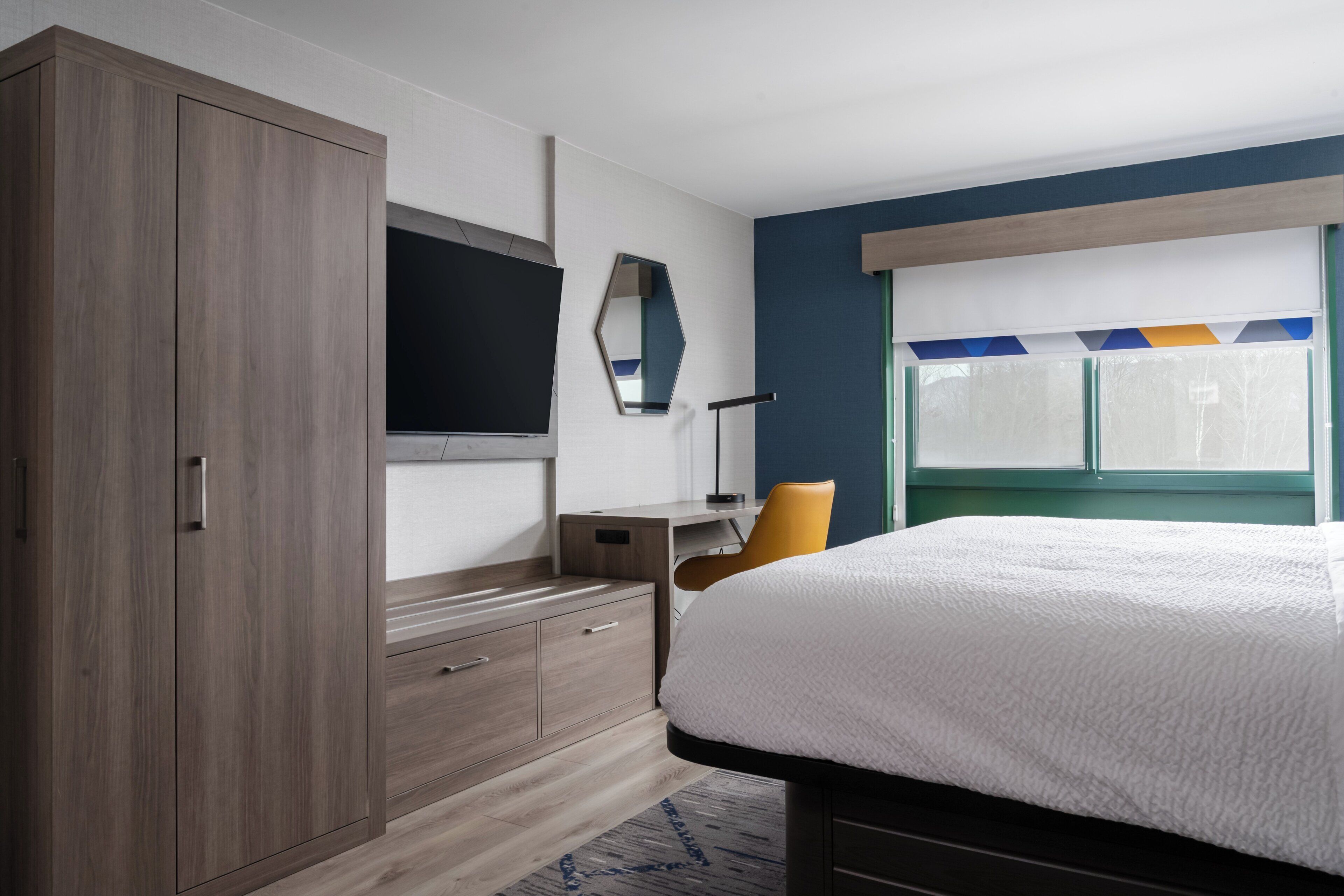 Ett modernt hotellrum med en stor säng, en garderob, ett skrivbord med stol, en platt-TV och ett fönster med utsikt över träd.