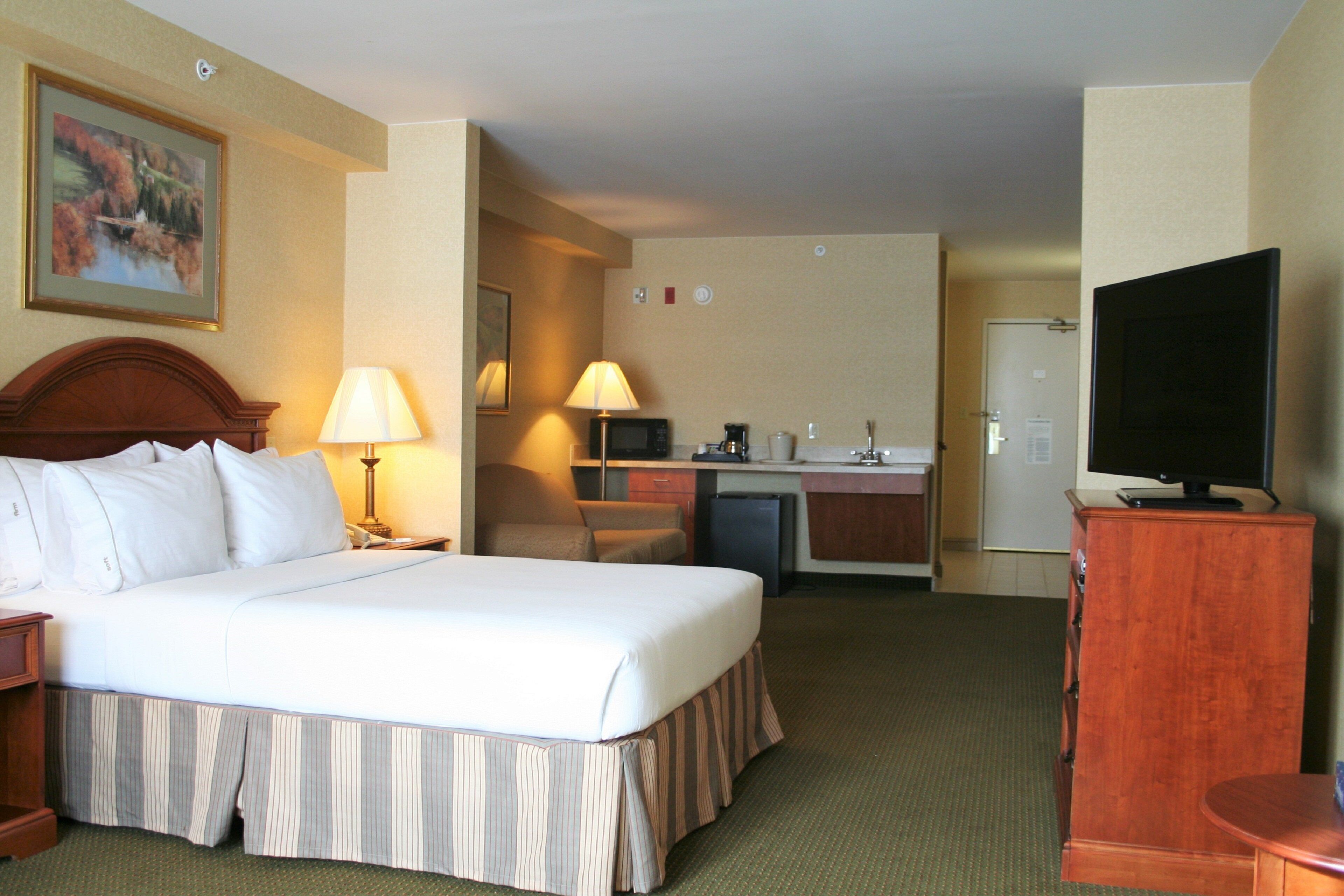 Habitación de hotel con cama, lámparas de noche, televisor, escritorio con computadora y una pequeña cocina.