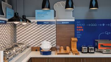 A breakfast station with labeled sections for fruit, bakery, and a pan-cake machine.