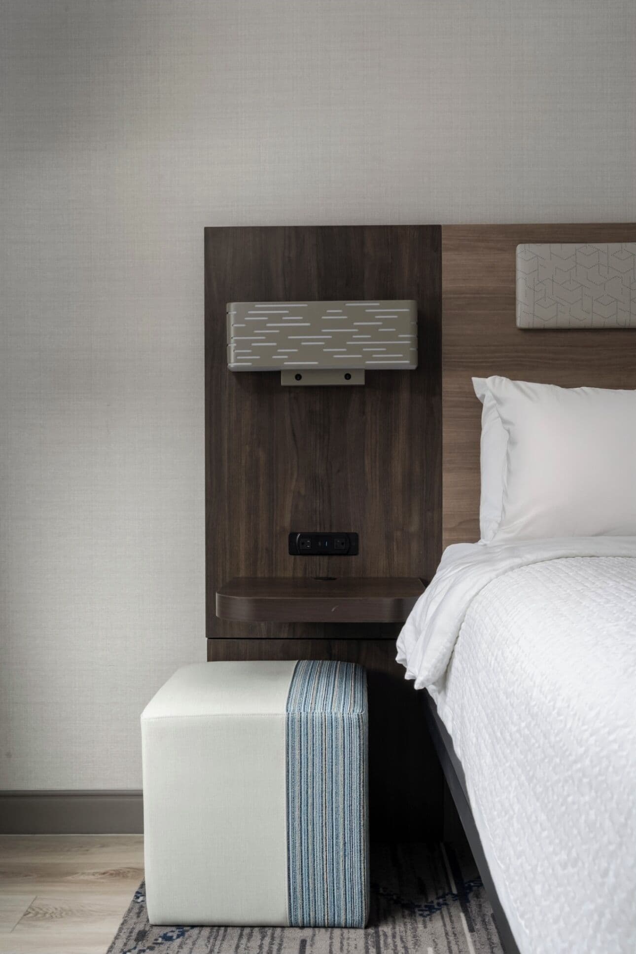 A modern hotel room with a wooden headboard, a bedside table with a power outlet, a white pillow, and a striped ottoman.
