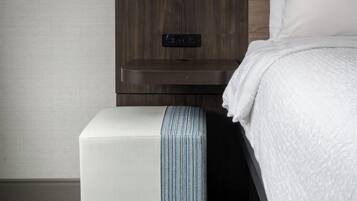 A modern hotel room with a wooden headboard, a bedside table with a power outlet, a white pillow, and a striped ottoman.