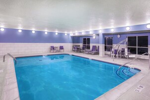 An indoor swimming pool with clear blue water, surrounded by purple lounge chairs and a tiled wall.