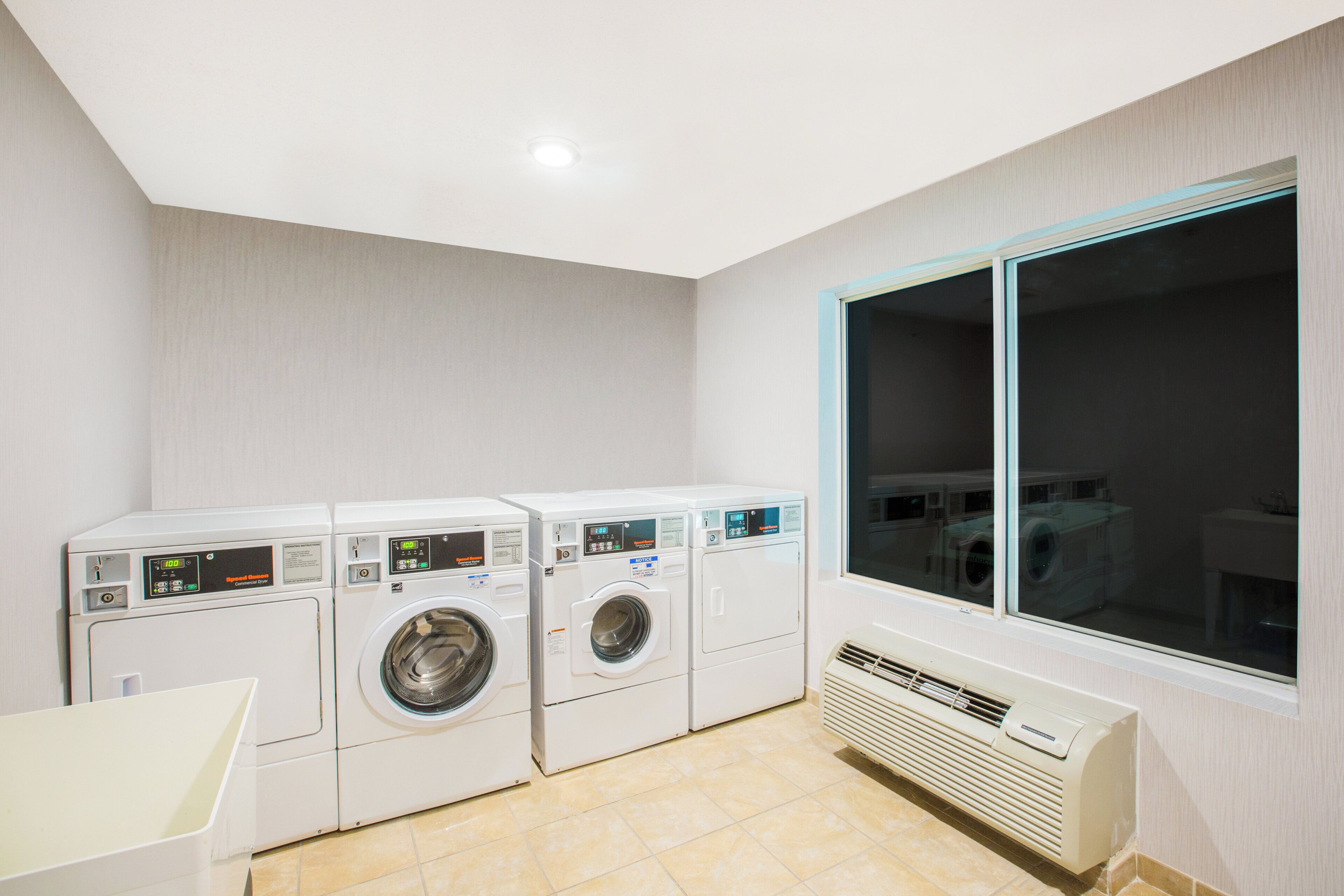 A laundry room with three stacked washing machines, a window with blinds, and a built-in air conditioning unit.