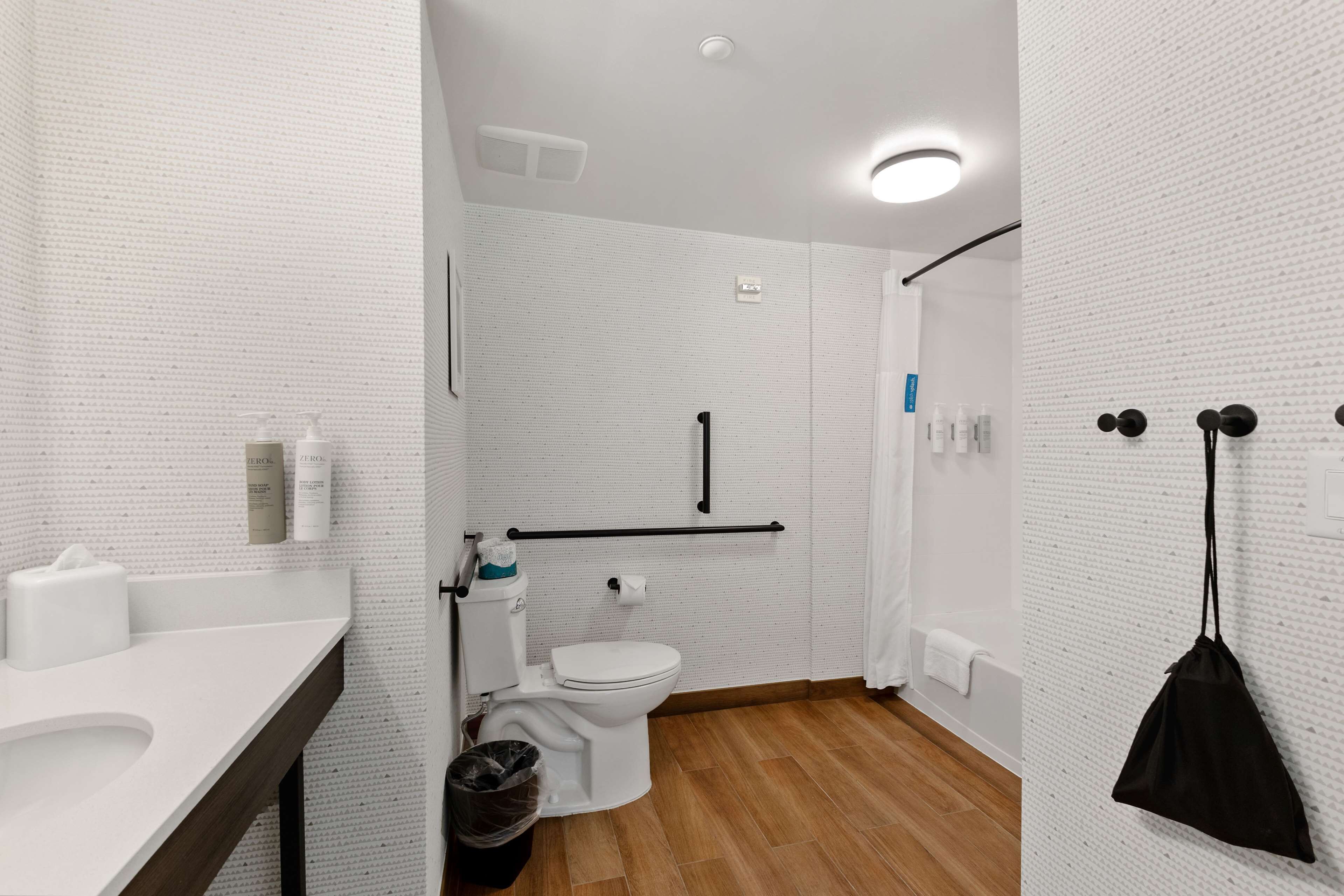 Une salle de bain moderne avec un mur recouvert de carreaux blancs, un plancher en bois, des toilettes blanches, un lavabo avec un miroir et un sac noir accroché au mur.