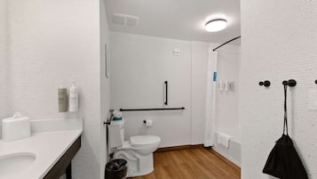 Une salle de bain moderne avec un mur recouvert de carreaux blancs, un plancher en bois, des toilettes blanches, un lavabo avec un miroir et un sac noir accroché au mur.