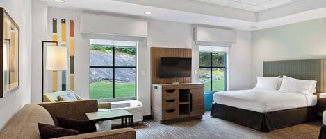 A modern hotel room with a large bed, a seating area with a sofa and armchair, a TV, and a view of the outdoors.