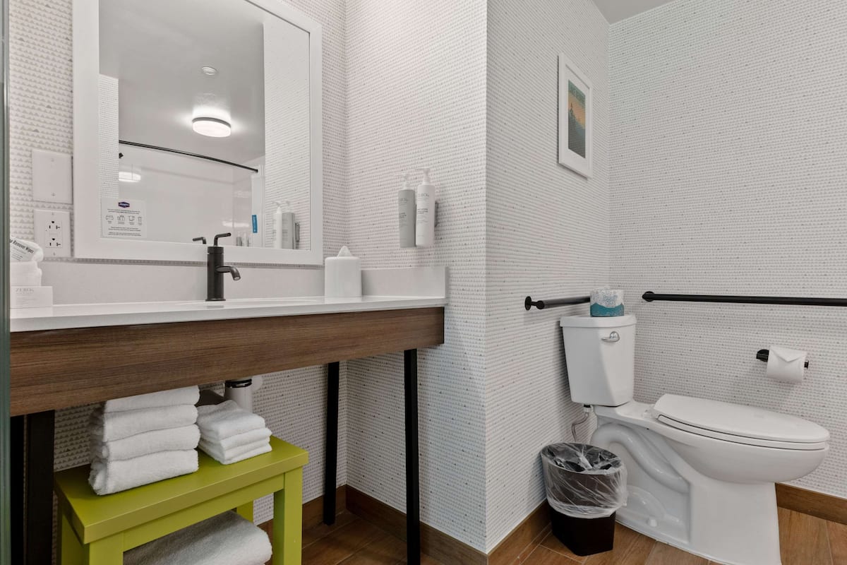 Um banheiro moderno com pia branca, espelho e vaso sanitário. Há um porta-toalhas verde e uma lixeira.