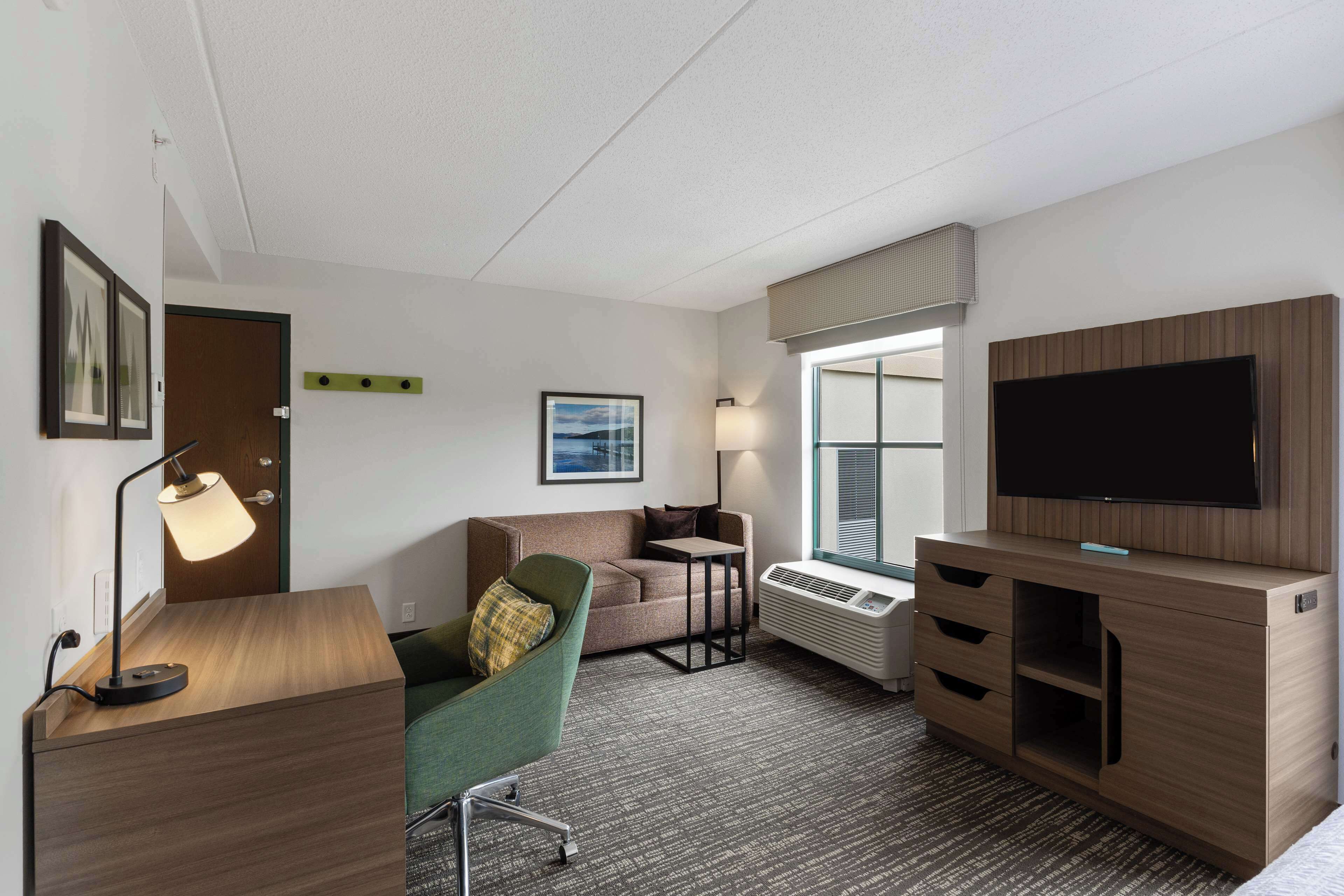 A hotel room with a desk, a green chair, a sofa, a flat-screen TV, a window with blinds, and a wall-mounted heater.