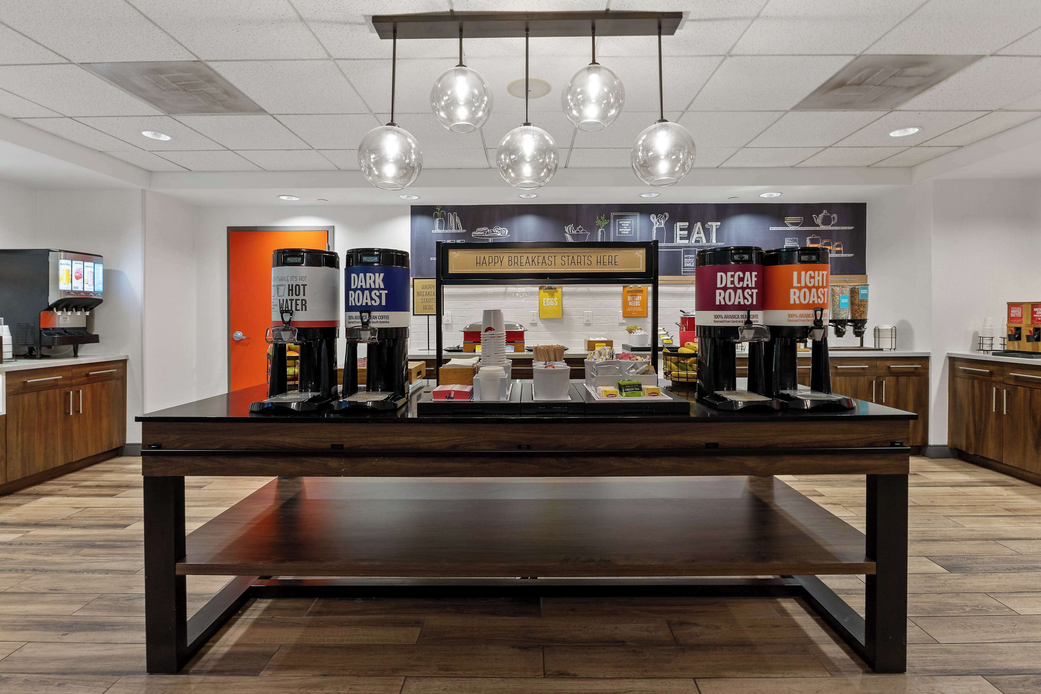 A breakfast area with a coffee machine, hot water dispenser, and a sign that says "HAPPY BREAKFAST STARTS HERE".