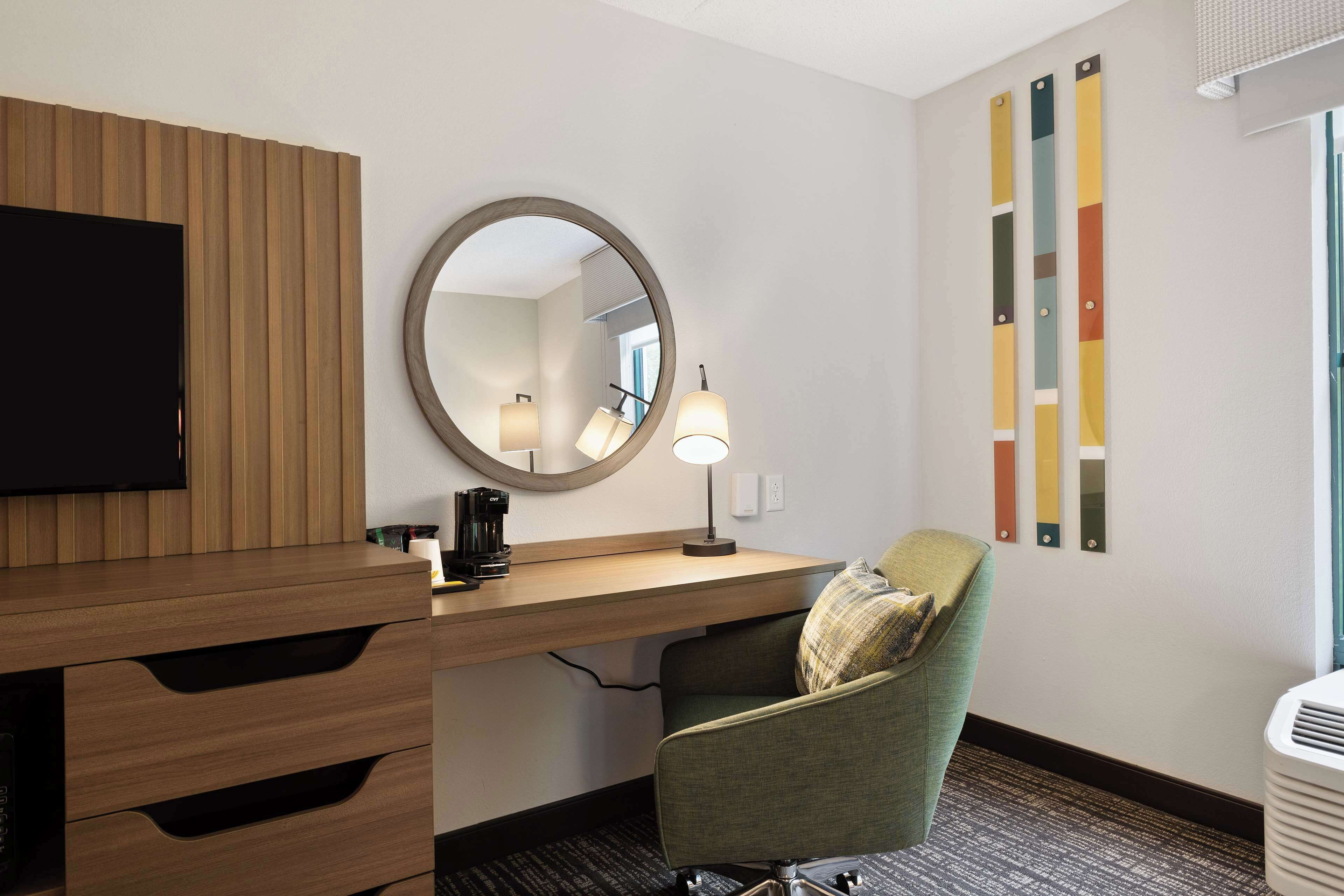 A hotel room with a wooden desk, a round mirror, a green armchair, a flat-screen TV, and a wall-mounted air conditioner.