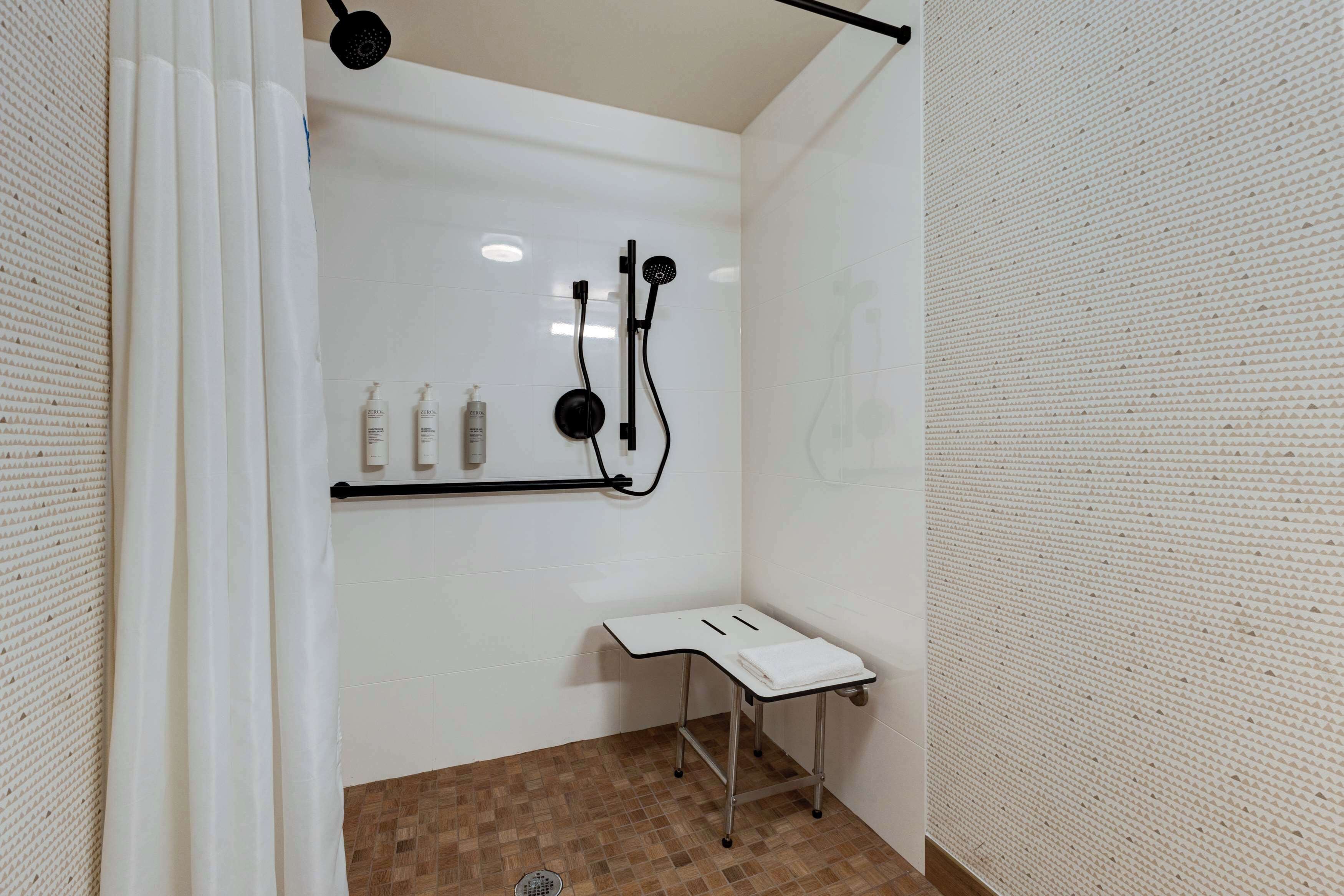 A shower area with a white tiled wall, a showerhead, a small table with a towel, and a drain.