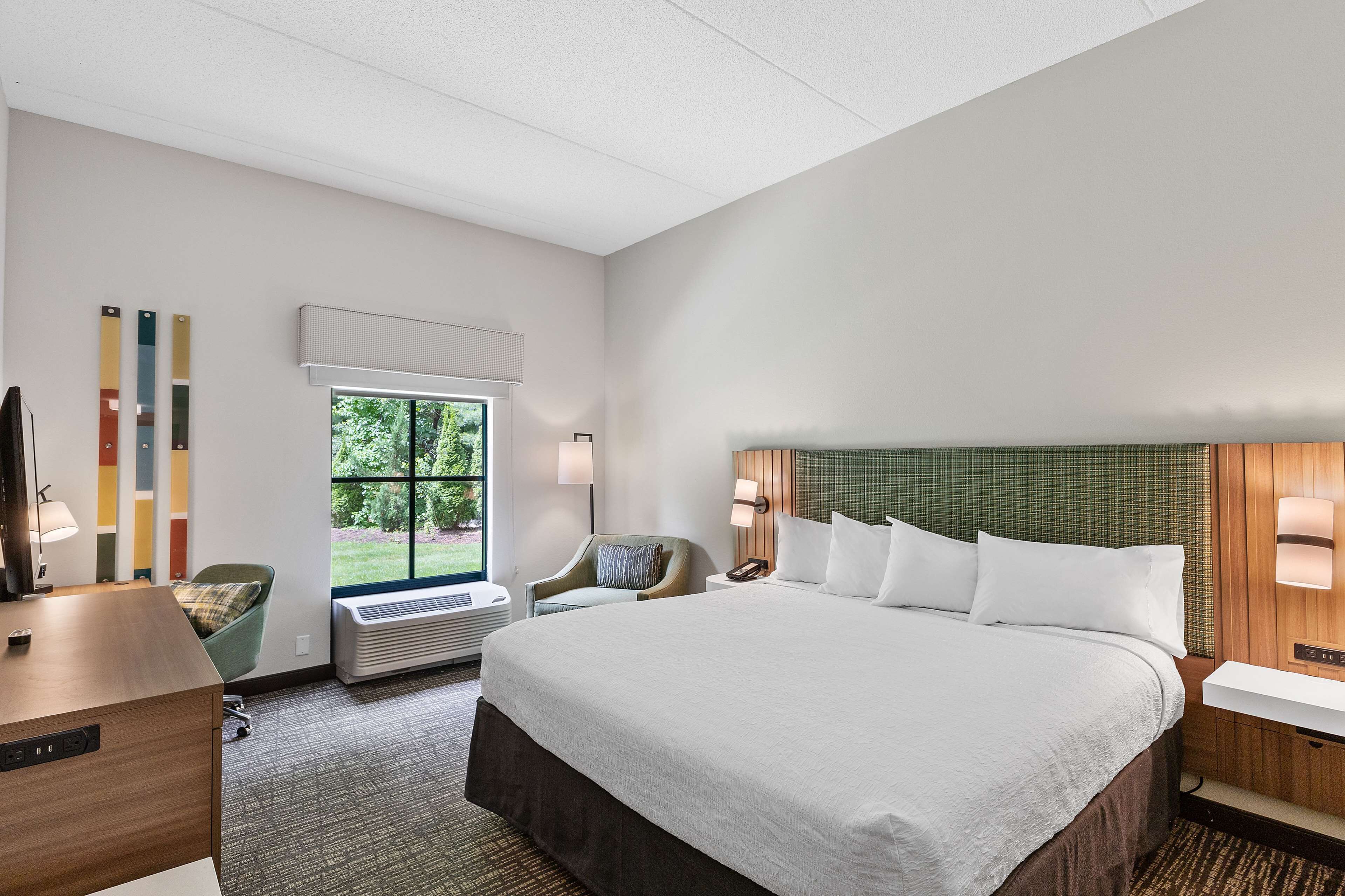 A hotel room with a large bed, a desk, a chair, a window with a view of greenery, and a wall-mounted air conditioner.