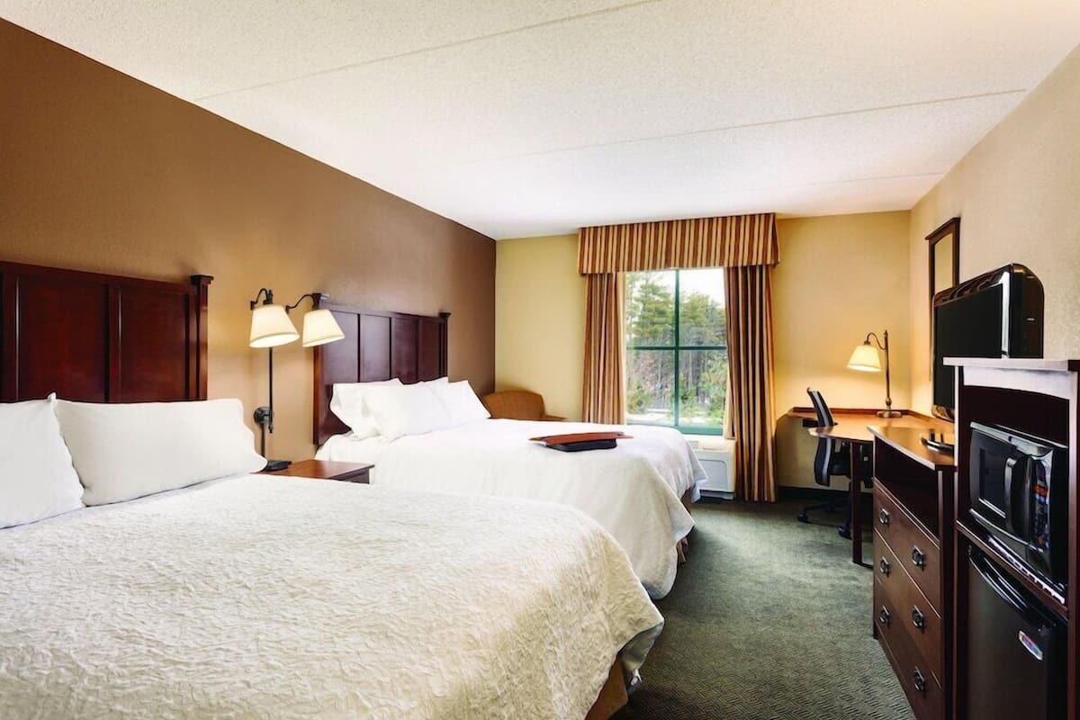 Quarto de hotel com duas camas, uma escrivaninha, cômoda, televisão e janela com cortinas.