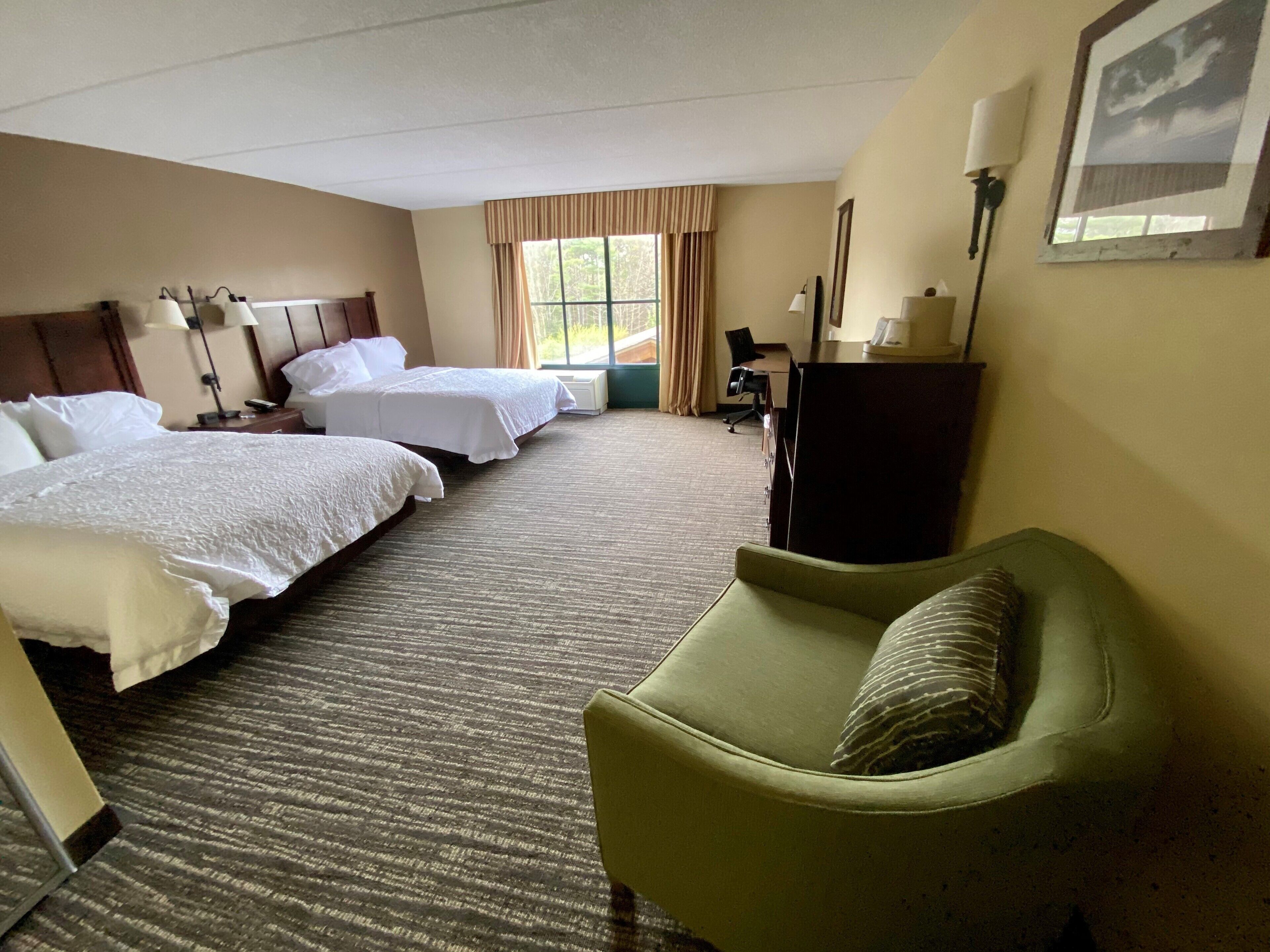 A hotel room with two beds, a green armchair, a desk, and a window with curtains.