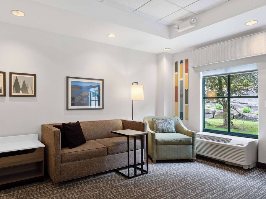 A modern hotel room with a sofa, armchair, and a small table. There is a window with a view of greenery outside.