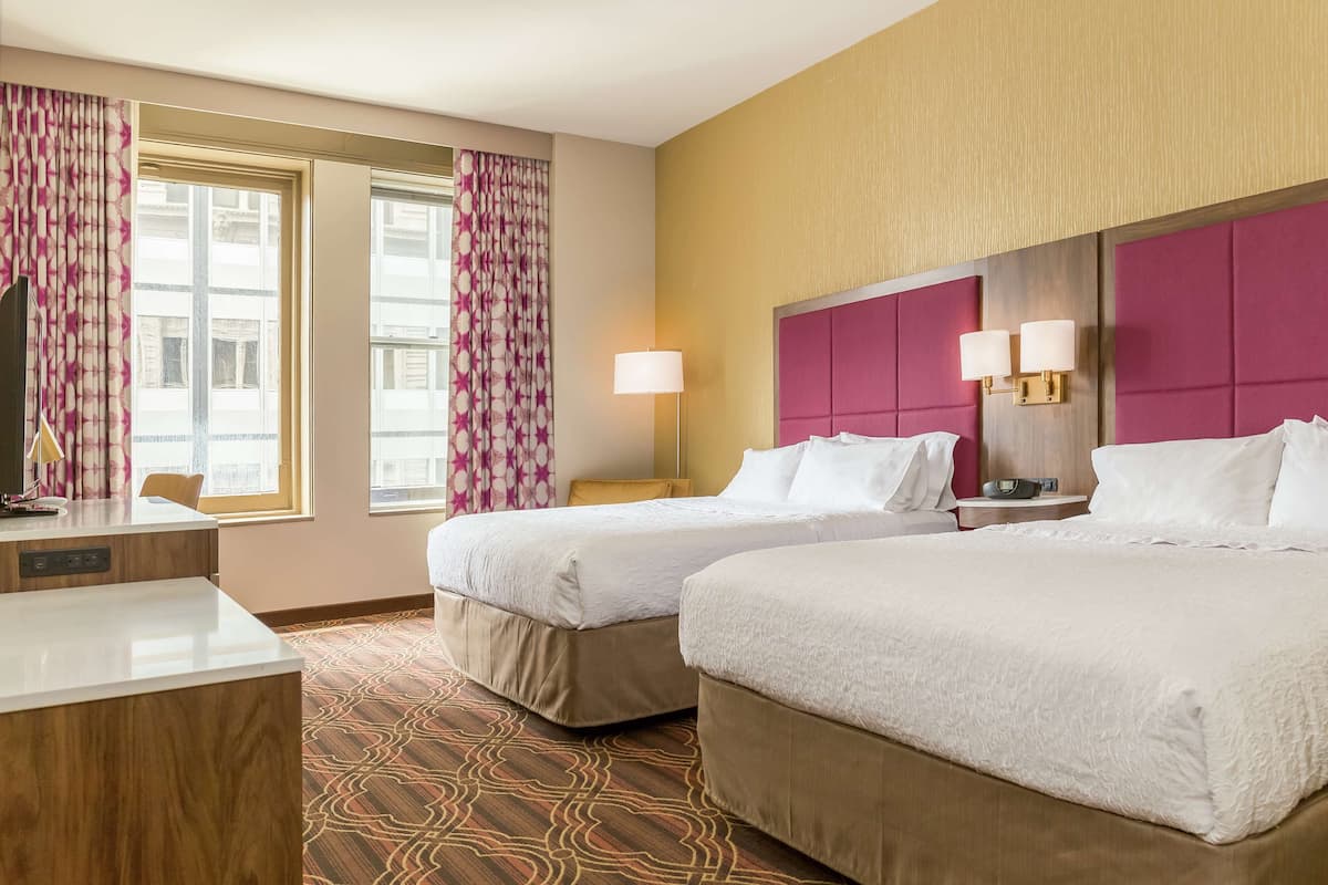 A hotel room with two beds, a desk, a lamp, and a view of the city.