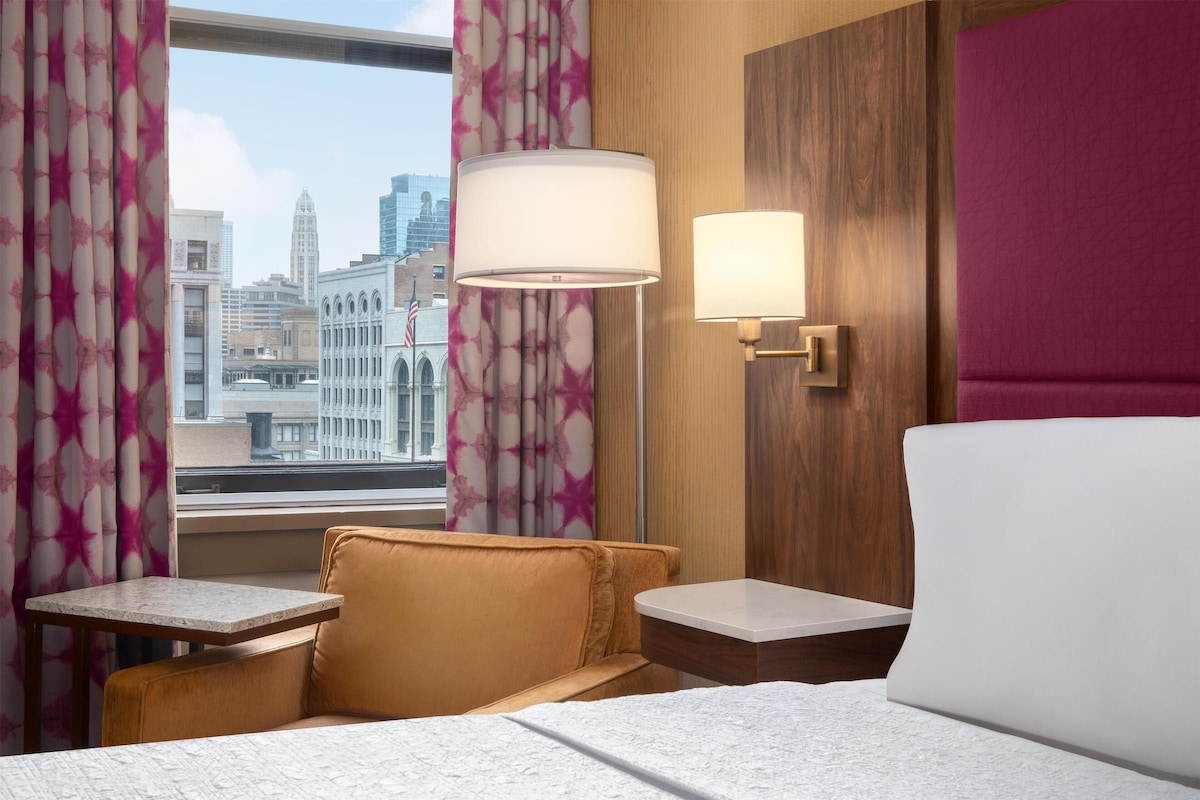 A hotel room with a bed, two bedside tables, a window with curtains, and a city view.