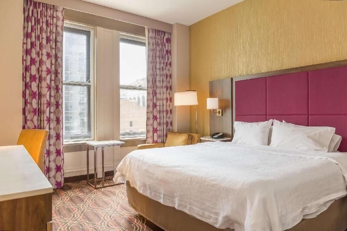 A hotel room with a bed, a desk, a chair, a window with curtains, and a nightstand with a lamp.